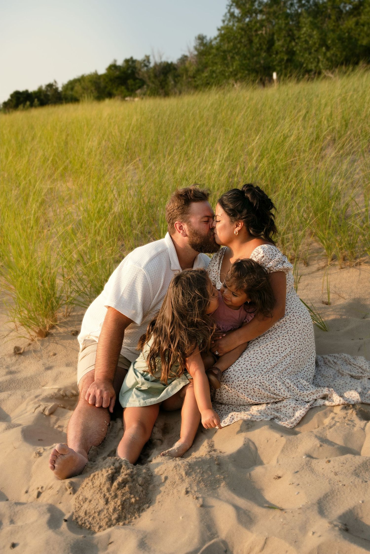 Happy family sharing a kiss on sandy beach dunes with lush green grass, parents embracing two young children.