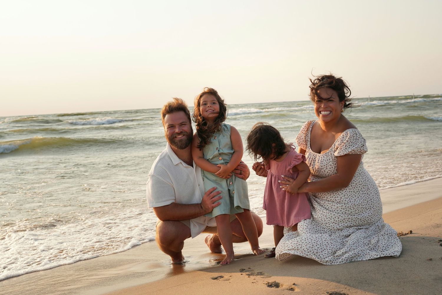 Happy family of four smiling together on a sandy beach at sunset, parents with two young daughters.