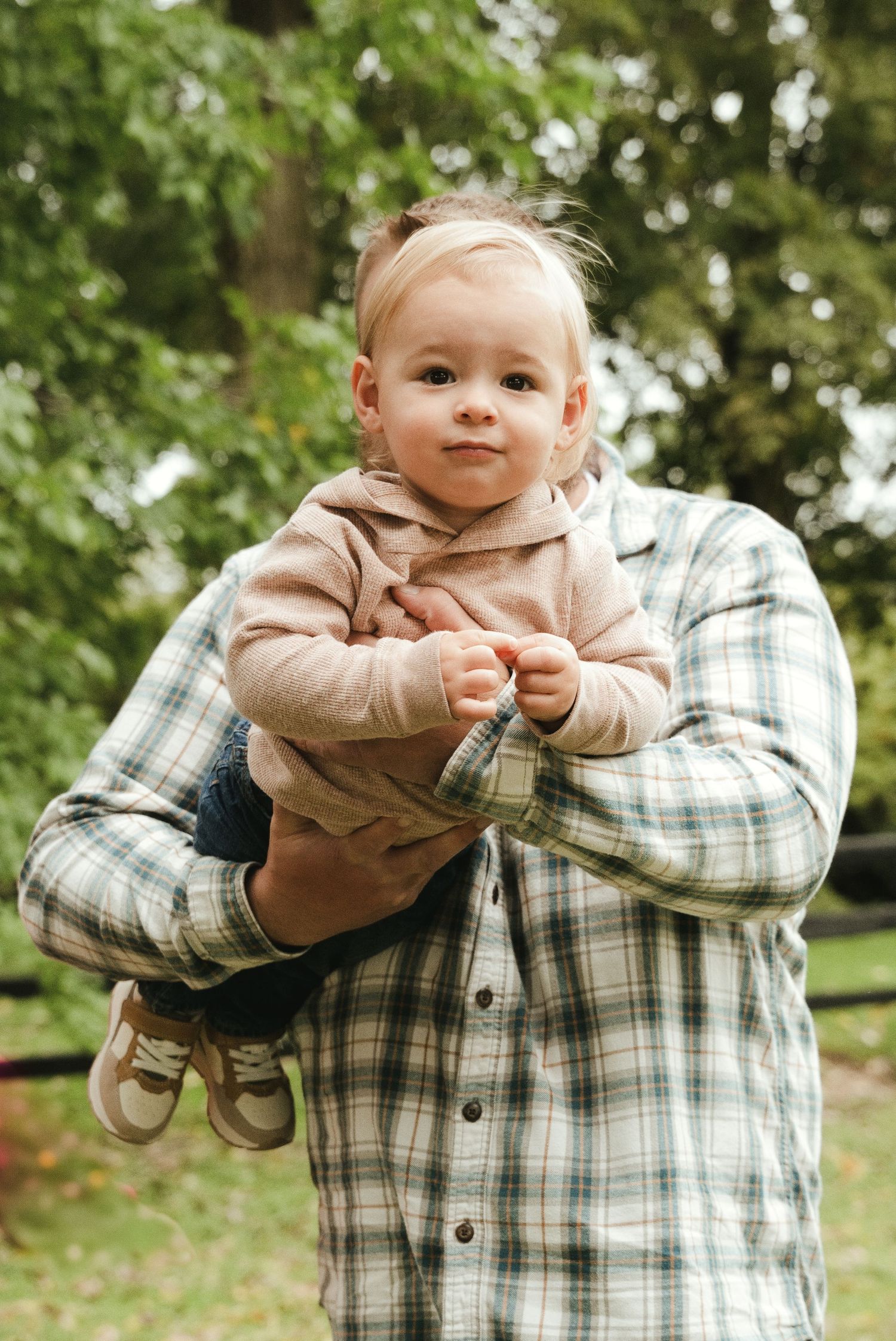 Smiling baby in a pink hooded sweater and sneakers held up in the air by a person in a plaid flannel shirt outdoors among green trees.