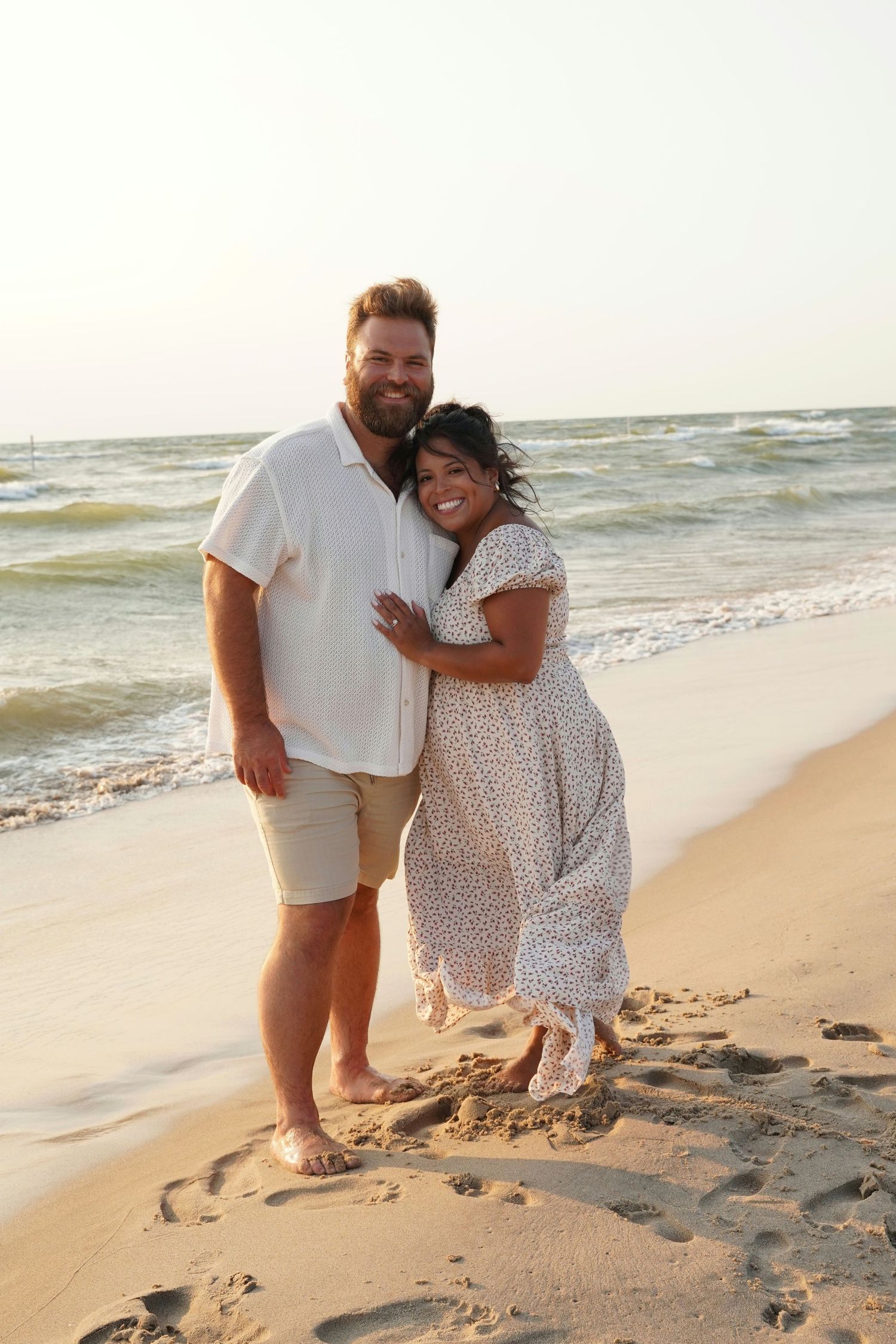 A happy couple embraces on a sandy beach at sunset, man in white shirt and shorts, woman in floral maxi dress.