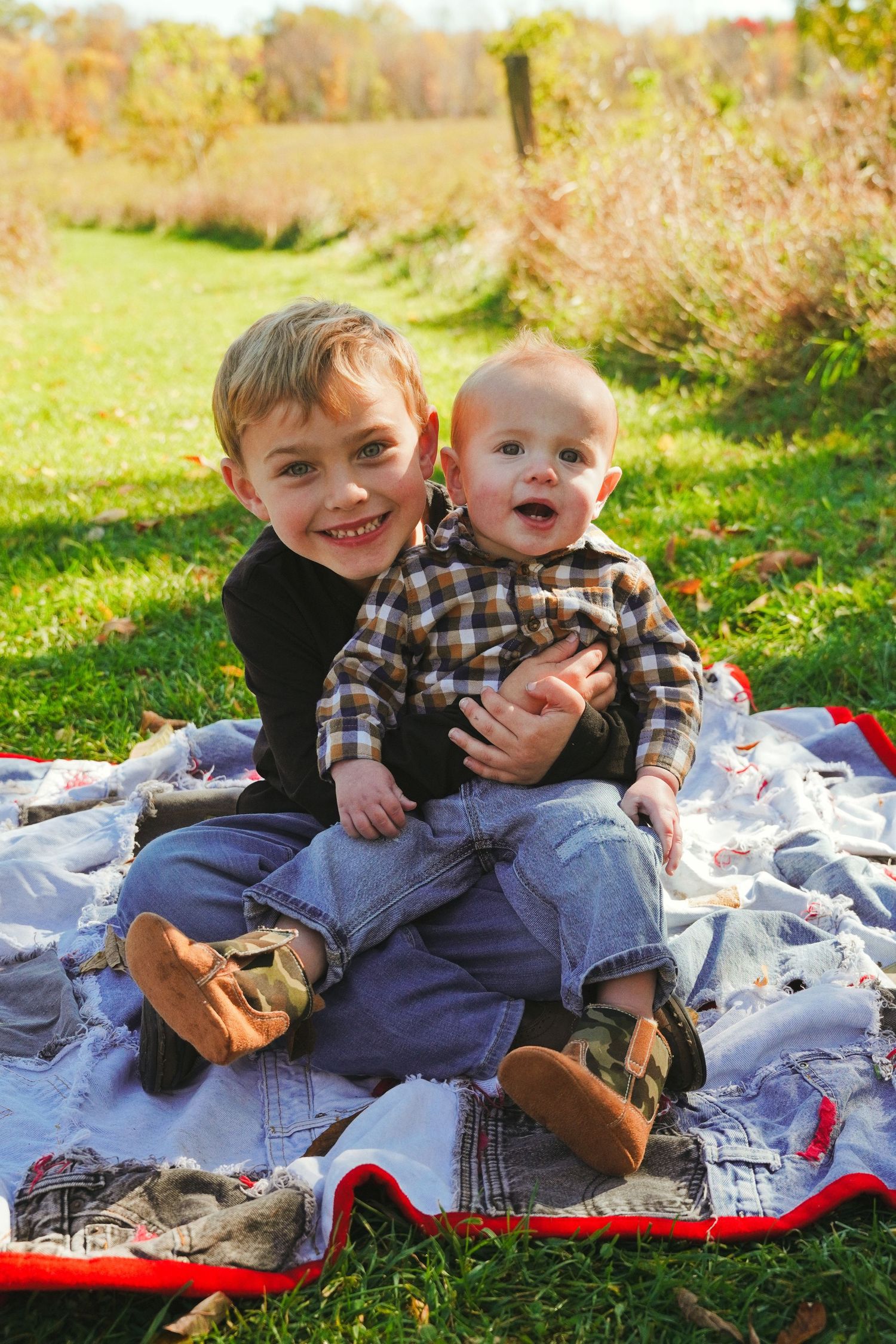Smiling older boy hugs a baby in a plaid shirt while sitting on a patchwork quilt on green grass in a sunny autumn field.