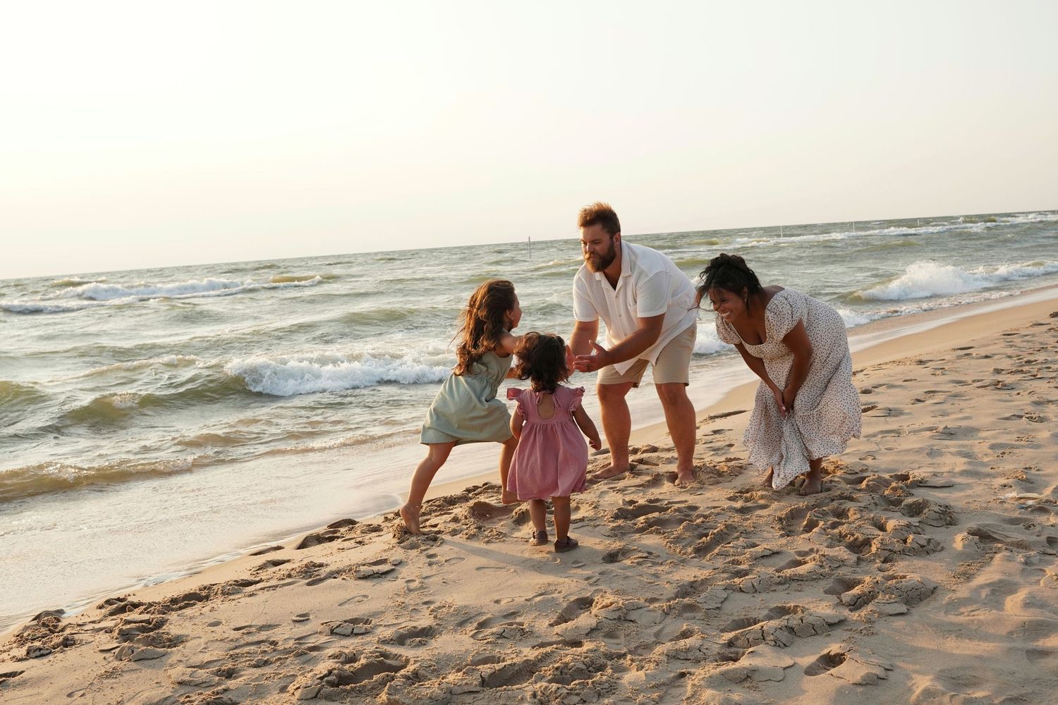 Family playing on sandy beach near ocean waves, parents holding hands with two young children on a sunny day.