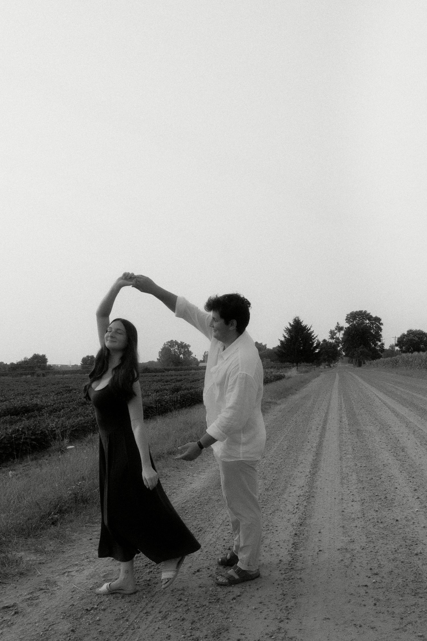 Black and white photo of couple dancing on a rural dirt road beside green farmland.