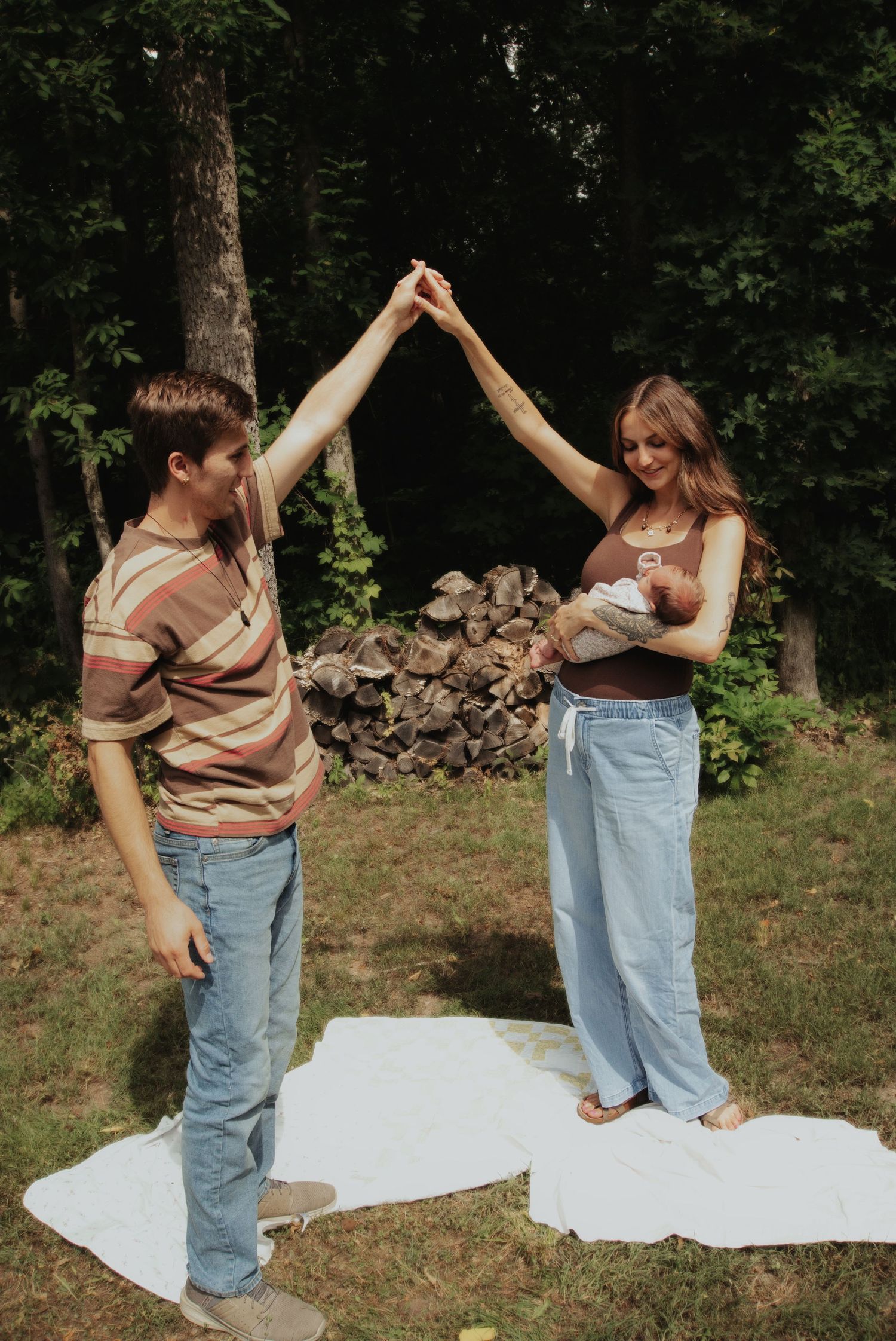 Young couple dancing outdoors holding hands overhead while mother cradles newborn baby in sunny backyard.