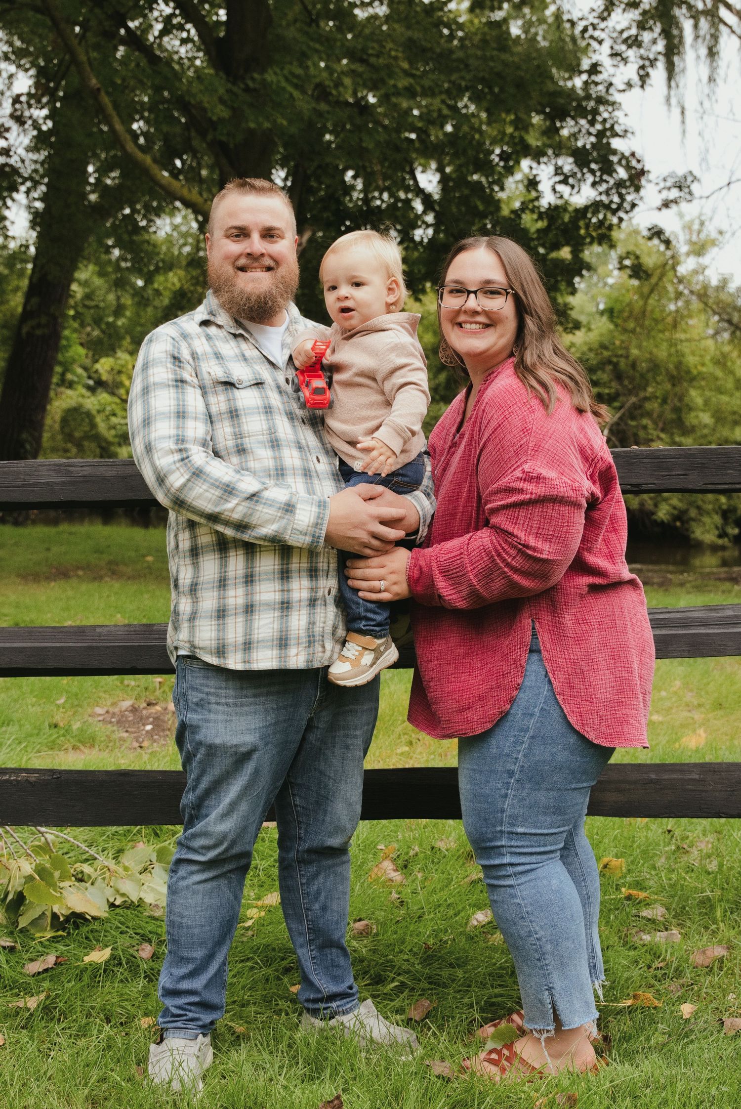 A smiling couple stands outdoors holding a toddler between them in front of a dark wooden fence, with green grass and trees behind.