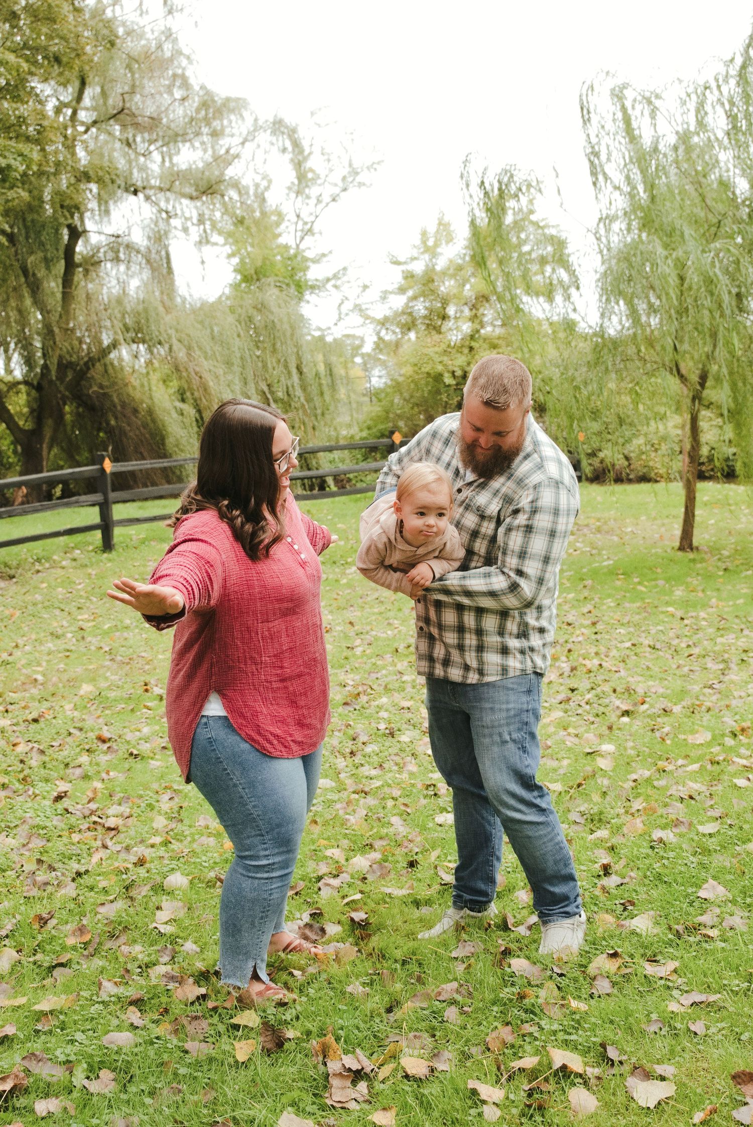 Smiling family of three playing on a grassy lawn with fallen leaves as a dad holds a baby beside a mom with arms outstretched, autumn.
