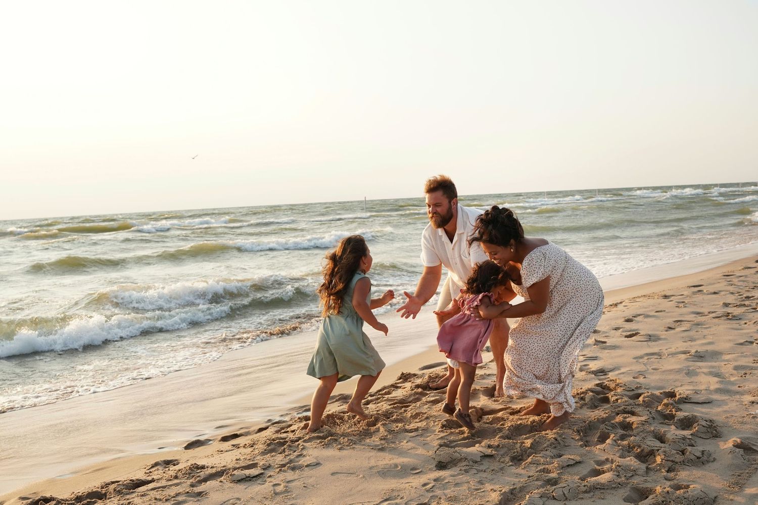 Family playing on a sunny beach, parents and two young girls dancing and laughing near the ocean waves.
