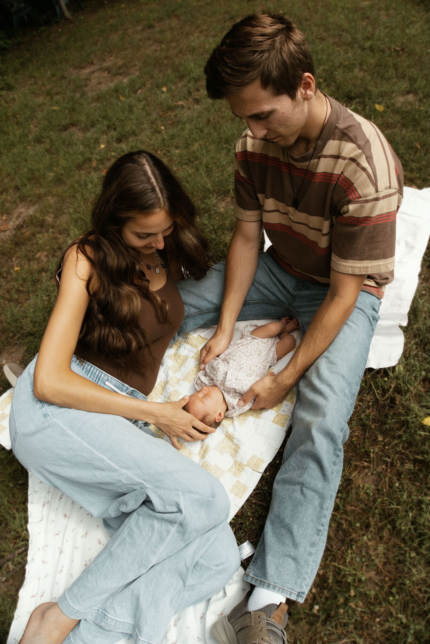 Young couple sitting on a blanket outdoors lovingly tending to their newborn baby.