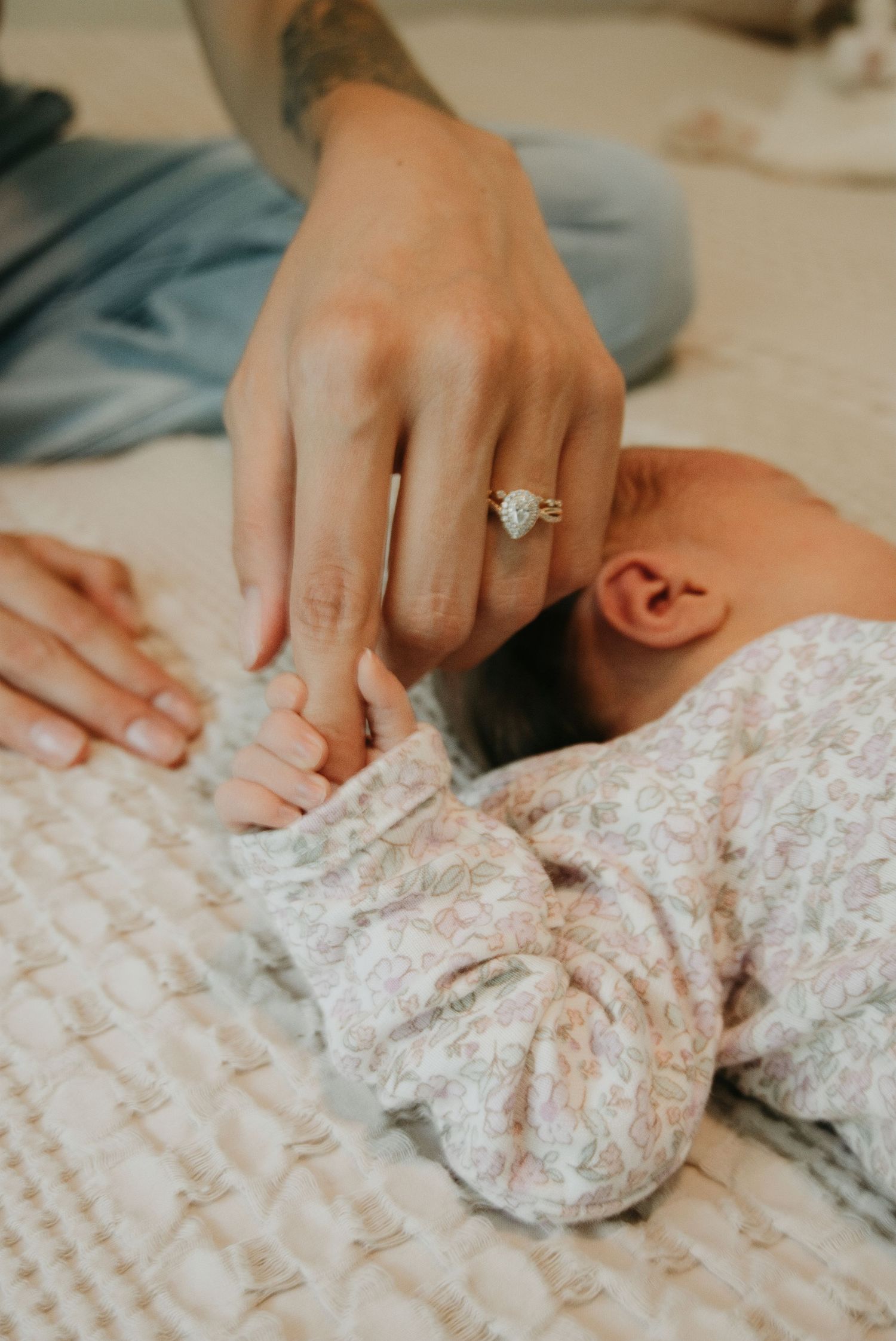 Close-up of a mother wearing a diamond engagement ring gently holding her newborn baby's hand on a soft quilted blanket.