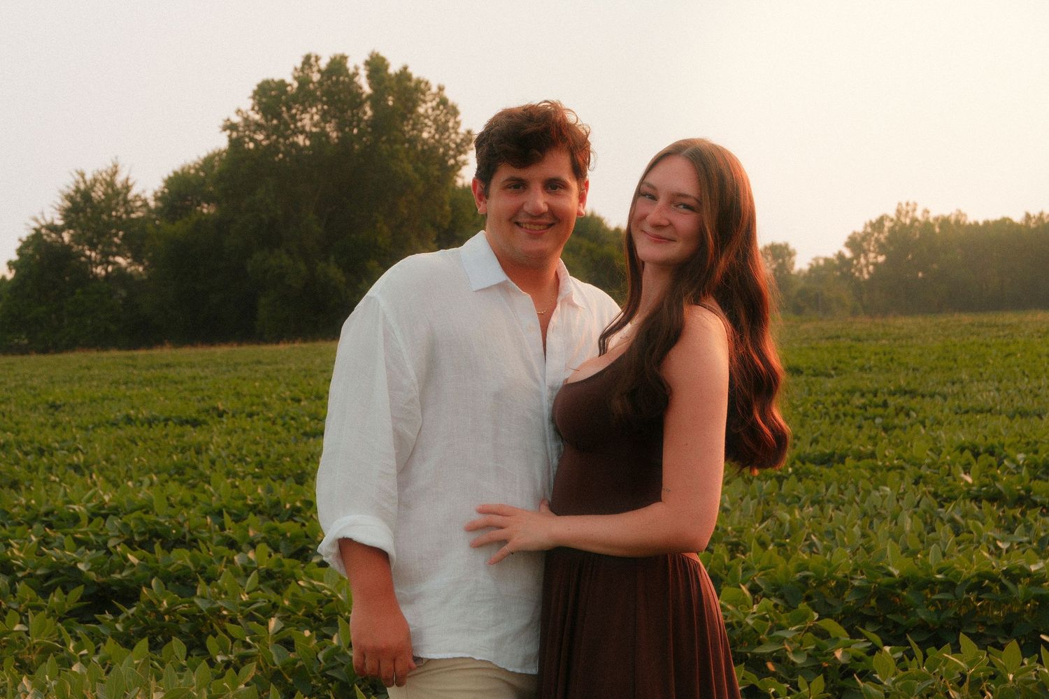 Couple posing together in a lush green field at sunset, woman in brown dress and man in white shirt.