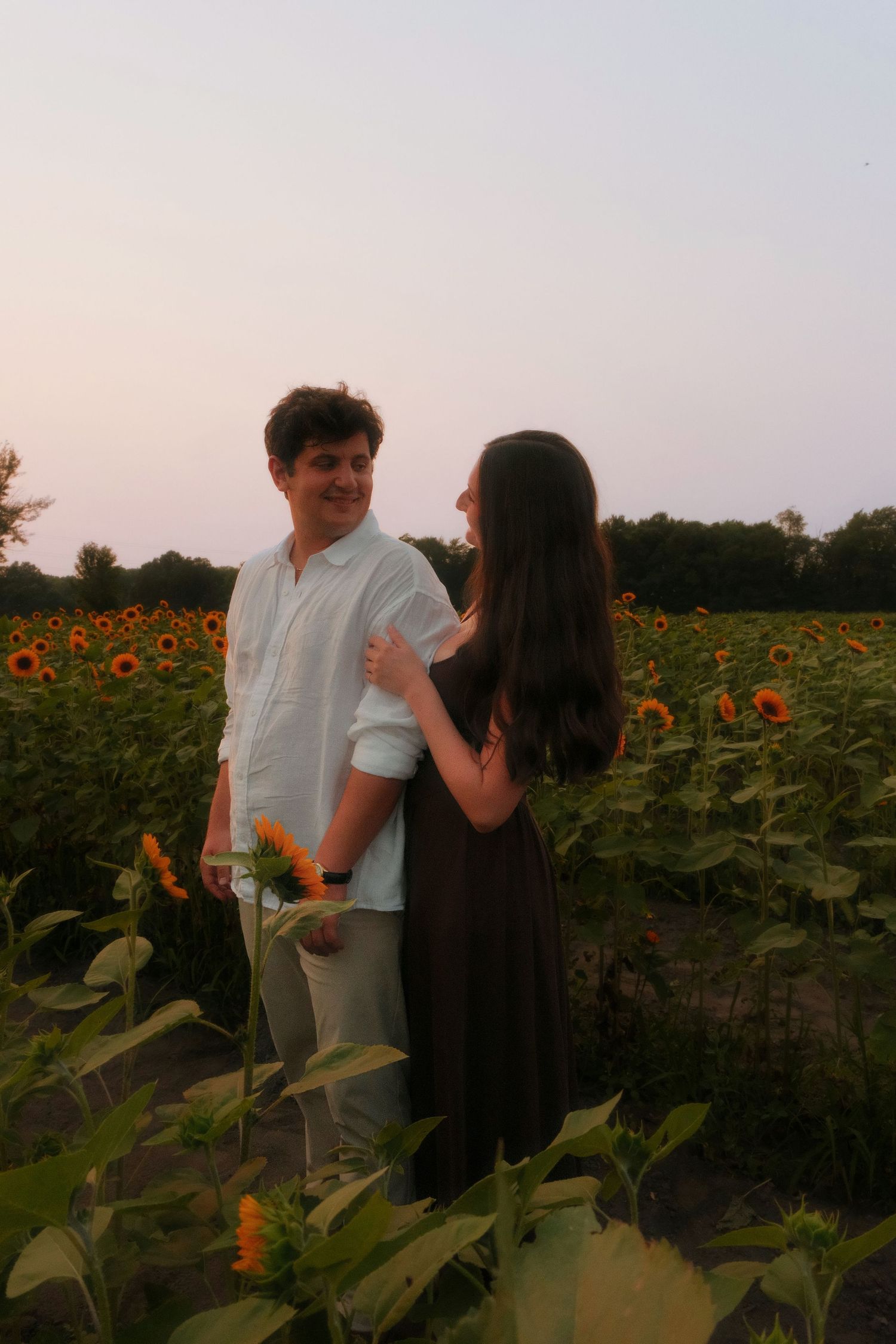 Couple sharing a romantic moment among tall sunflowers in a field during a soft pink sunset.