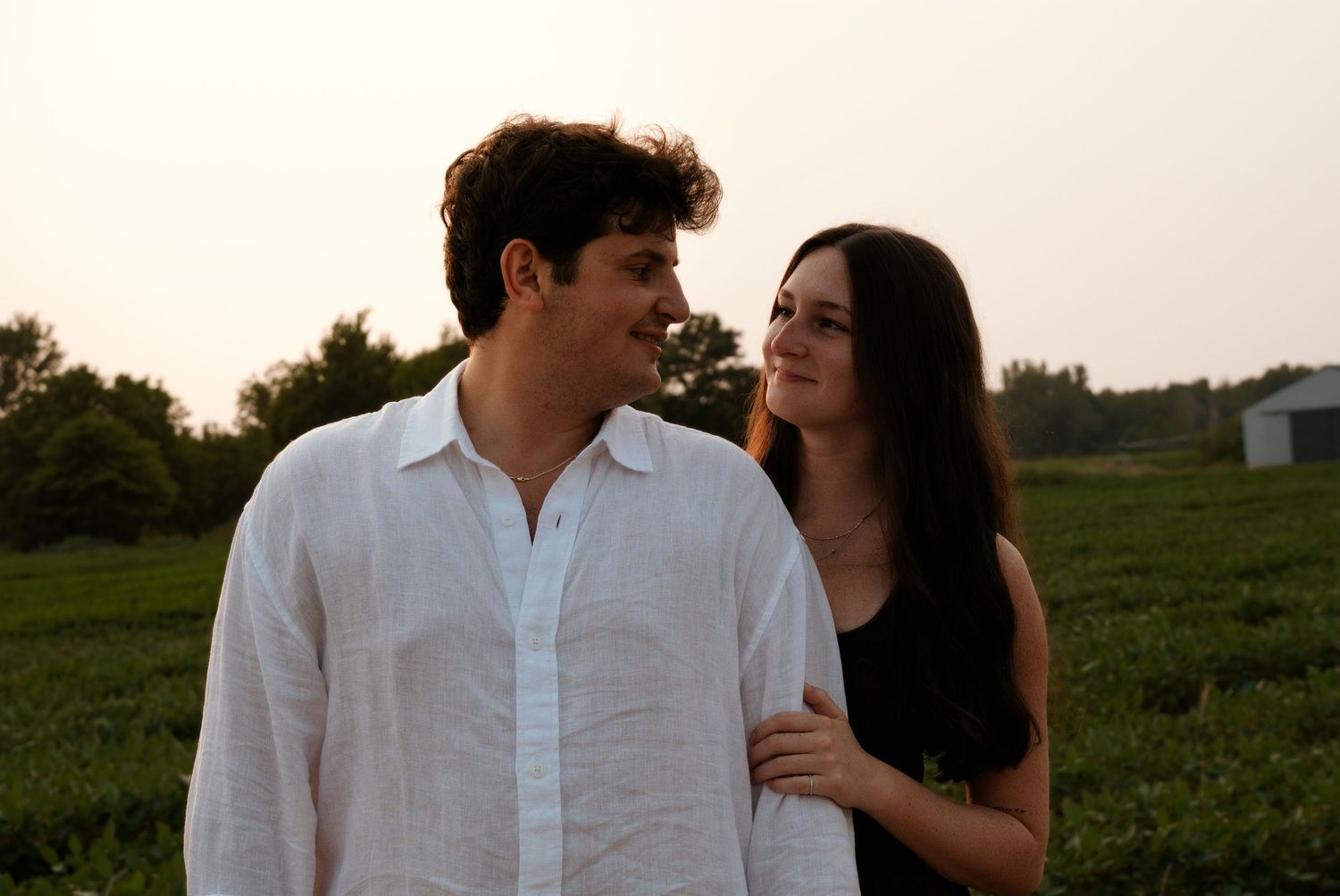 Young couple smiling at each other in front of a green field at sunset.