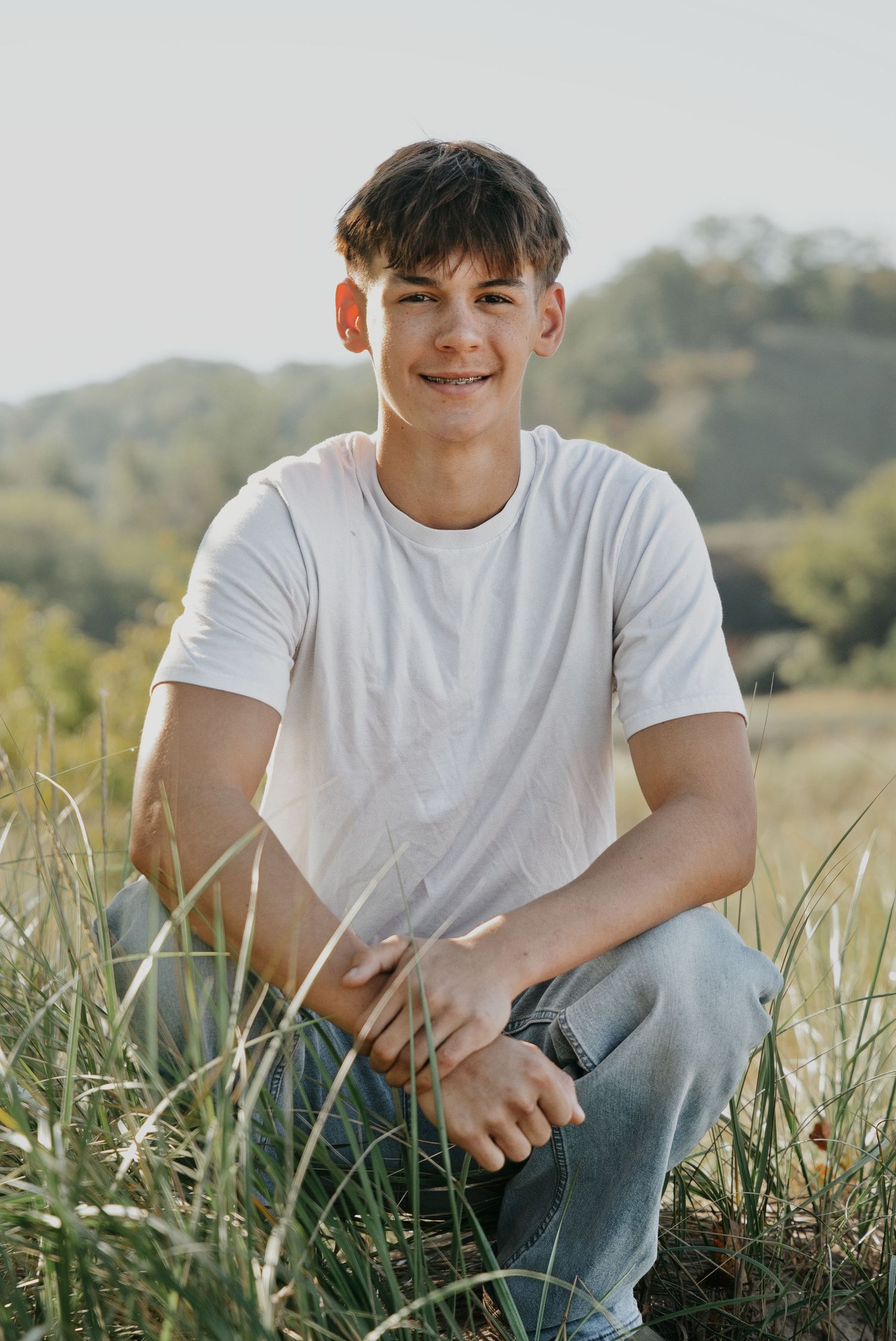 A teenage boy in a white t-shirt and jeans sits smiling in a grassy field with trees and hills behind him.