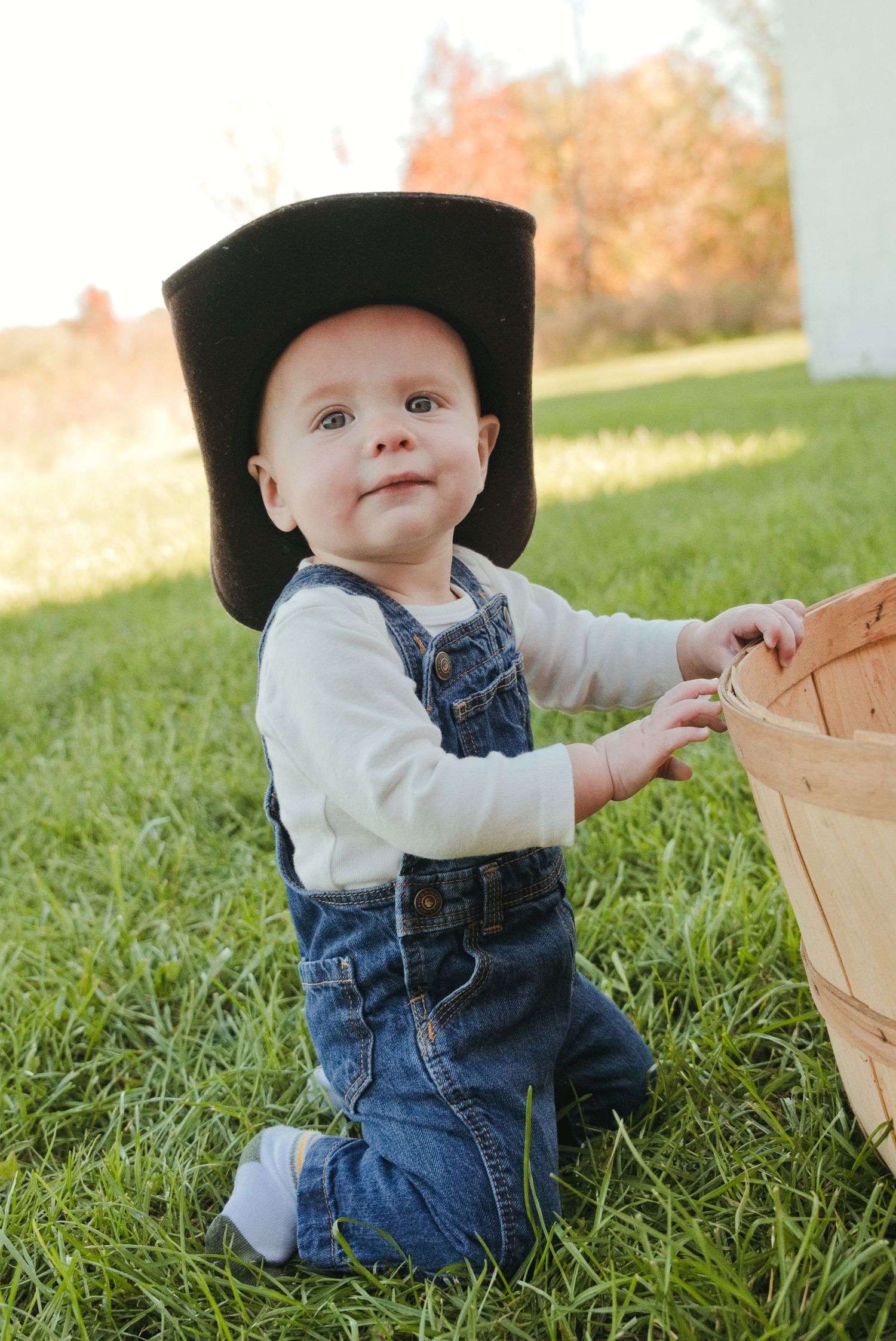 Baby in denim overalls and an oversized black hat kneels on green grass beside a wooden bushel basket on a sunny autumn day.