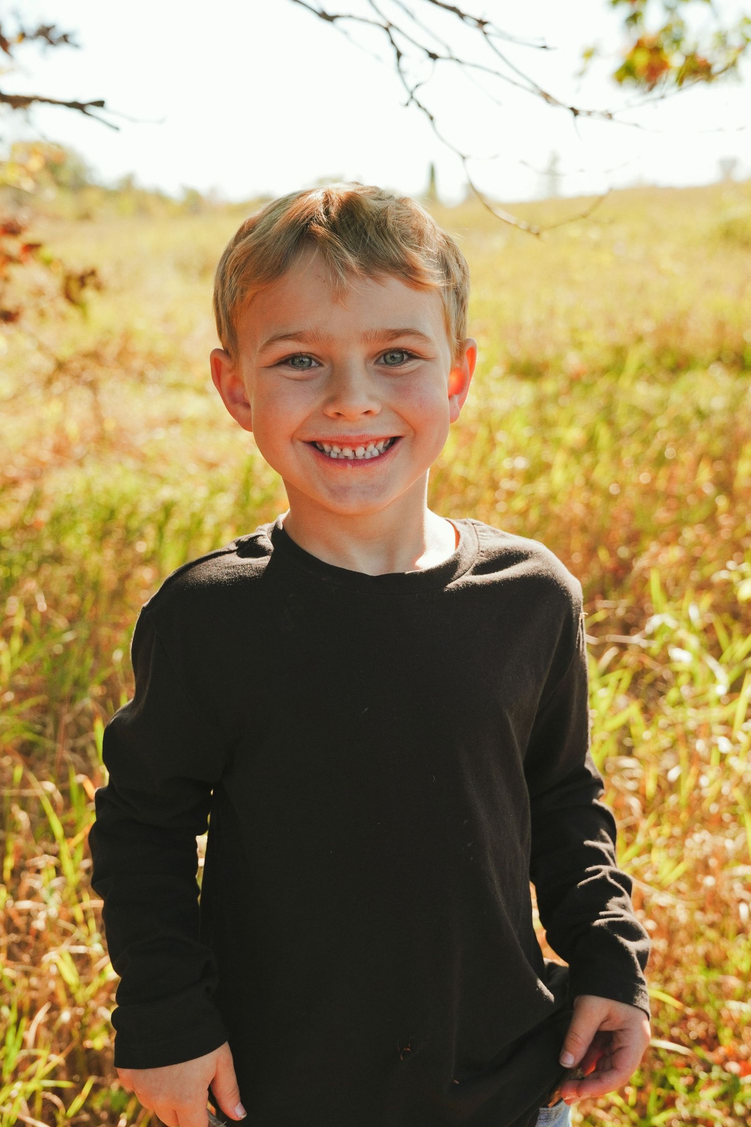 Smiling young boy in a black long-sleeve shirt standing in a sunlit grassy field with golden autumn foliage behind him.