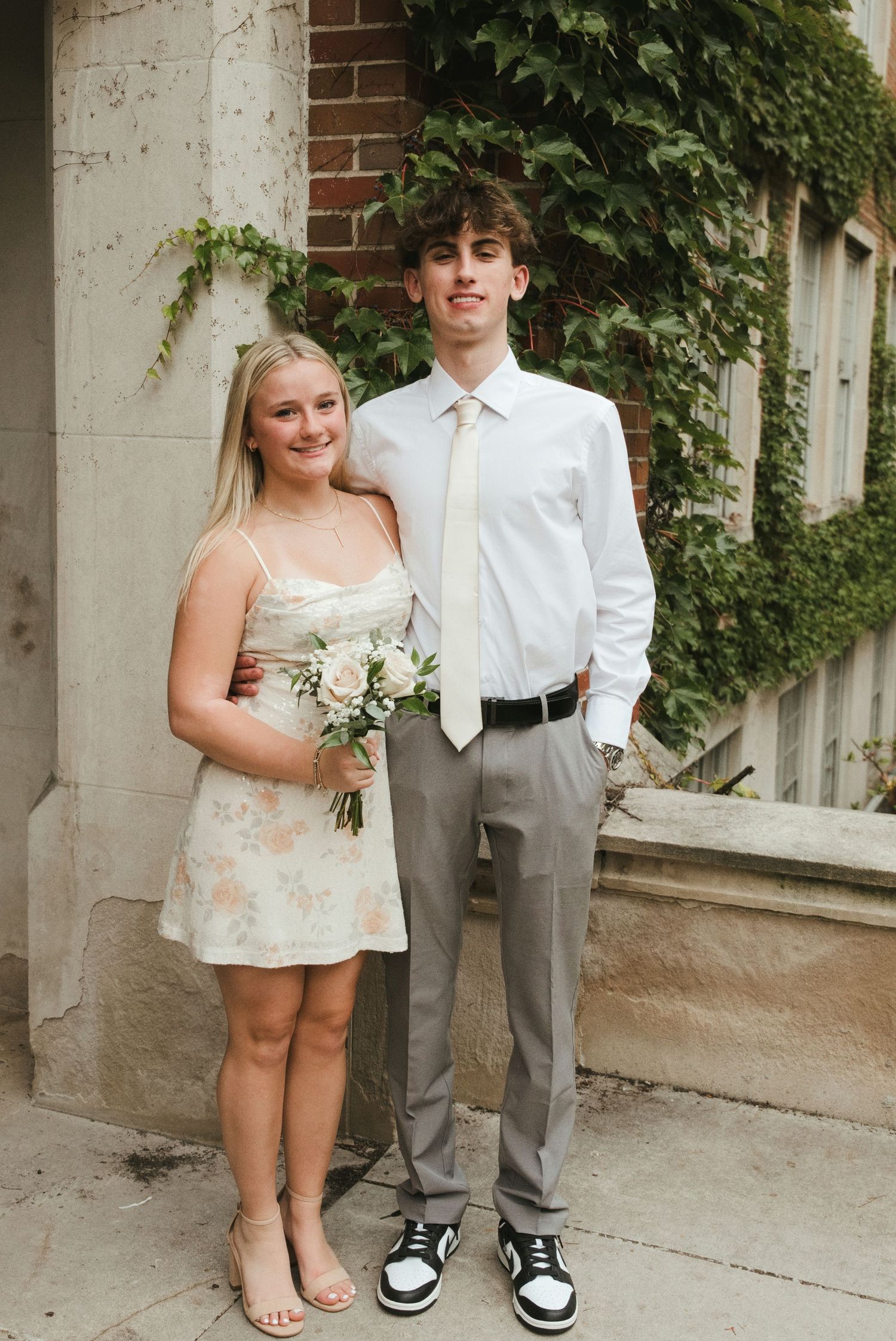 A smiling young couple poses outdoors by a brick wall with ivy, she in a floral sundress holding flowers, he in a white shirt and tie.