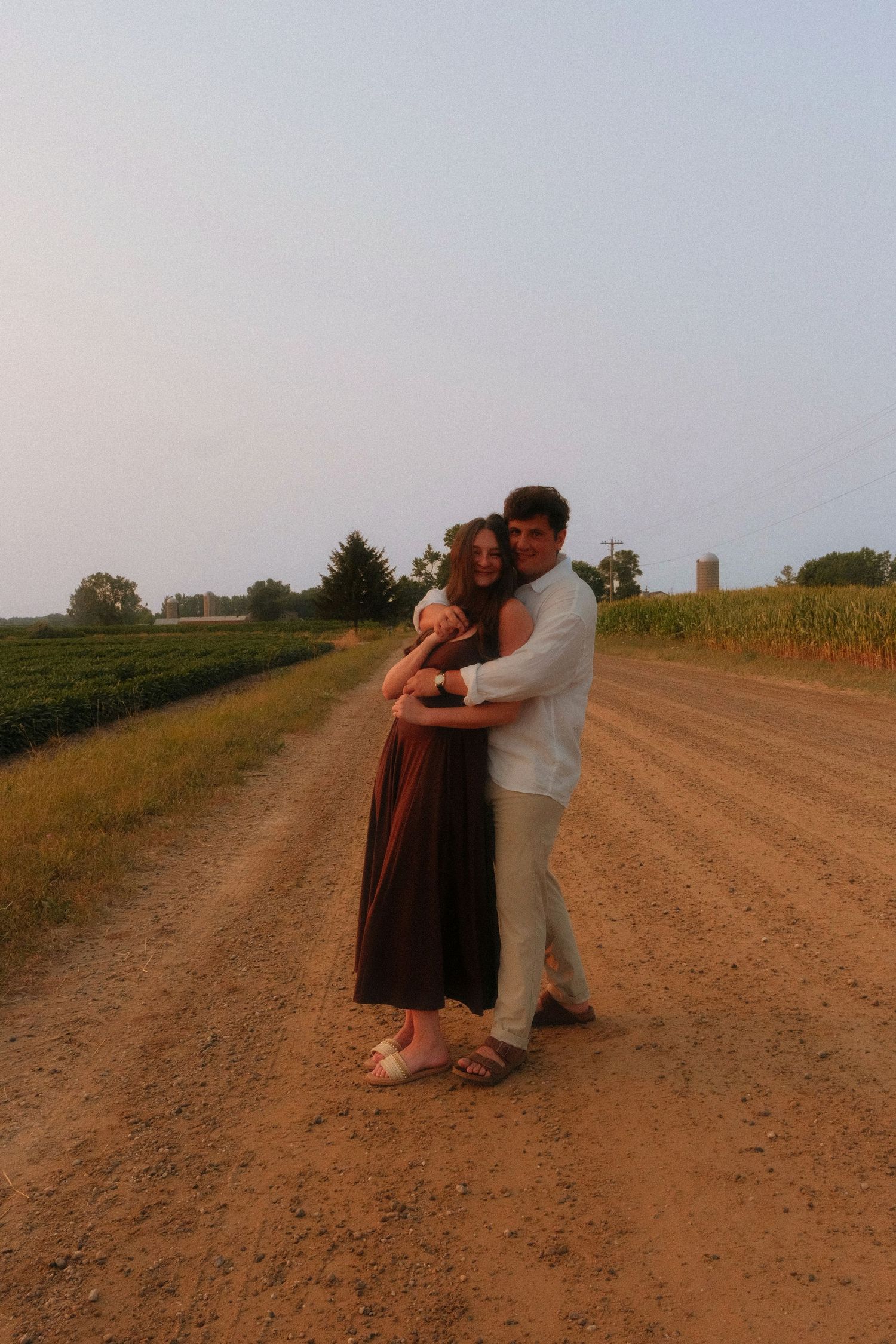 A happy couple embraces on a rustic dirt road surrounded by farm fields during golden hour sunset.