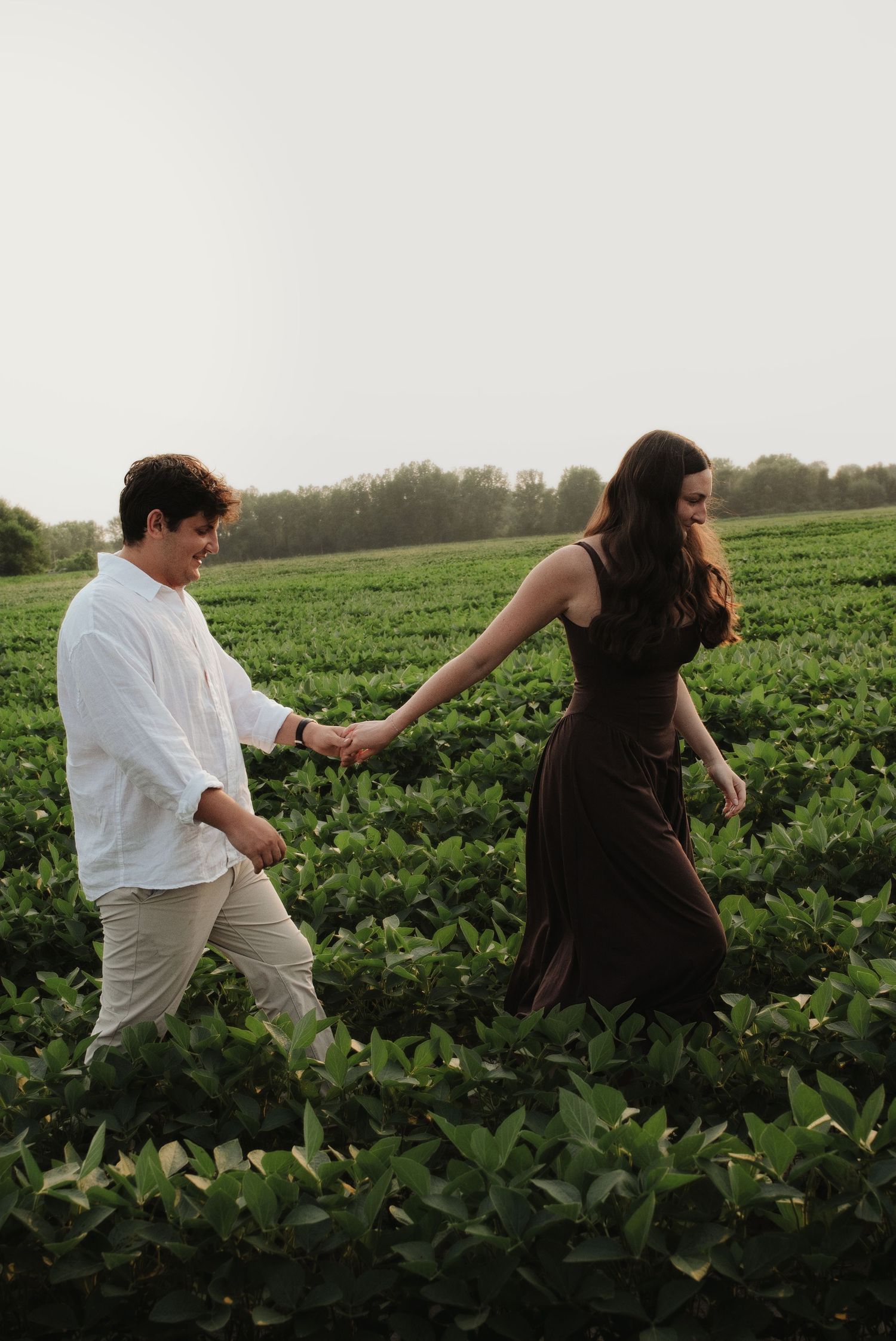 Couple holding hands and walking through a green field at dusk, woman in dark dress leads the way.