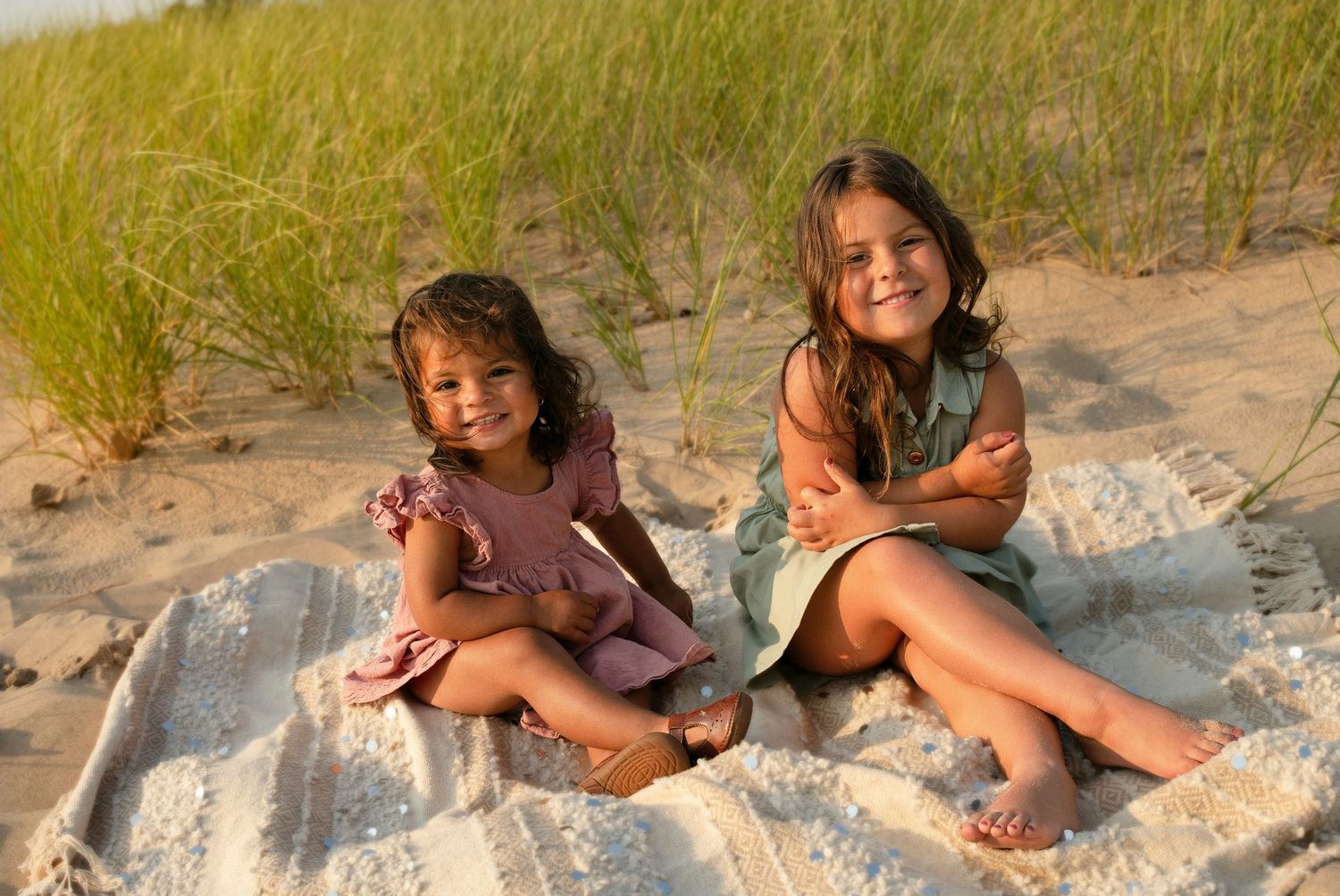 Two young girls sitting on a blanket on a sandy beach with green dunes, smiling in warm golden light.