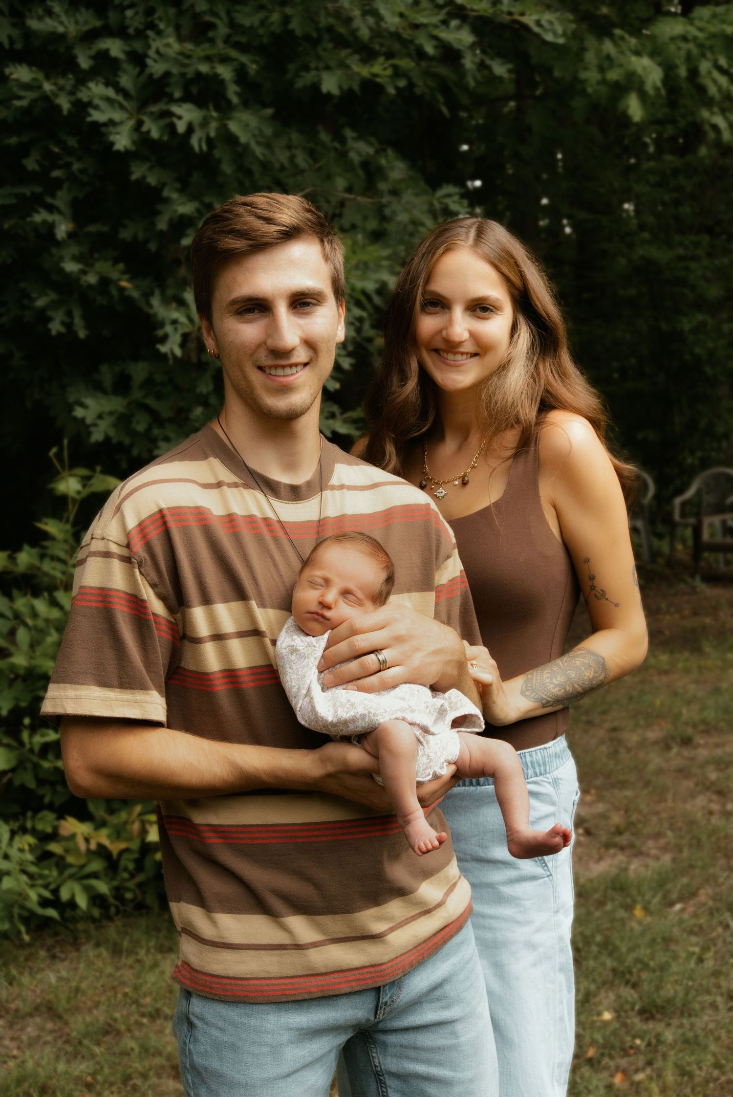 Young couple smiling outdoors holding their newborn baby wrapped in white lace clothing.