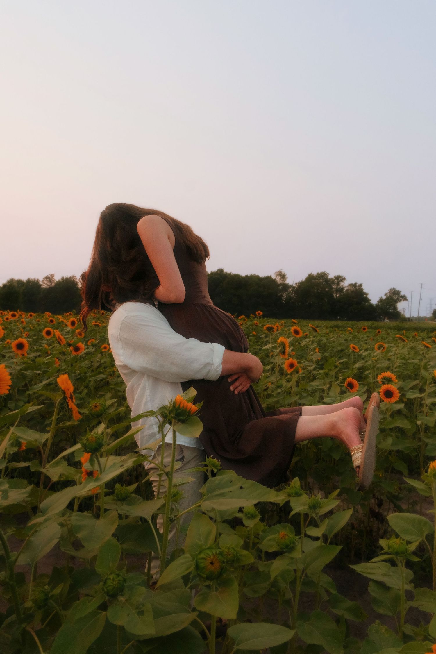 Man lifting woman in brown dress in a sunflower field at sunset, sharing a joyful romantic moment.