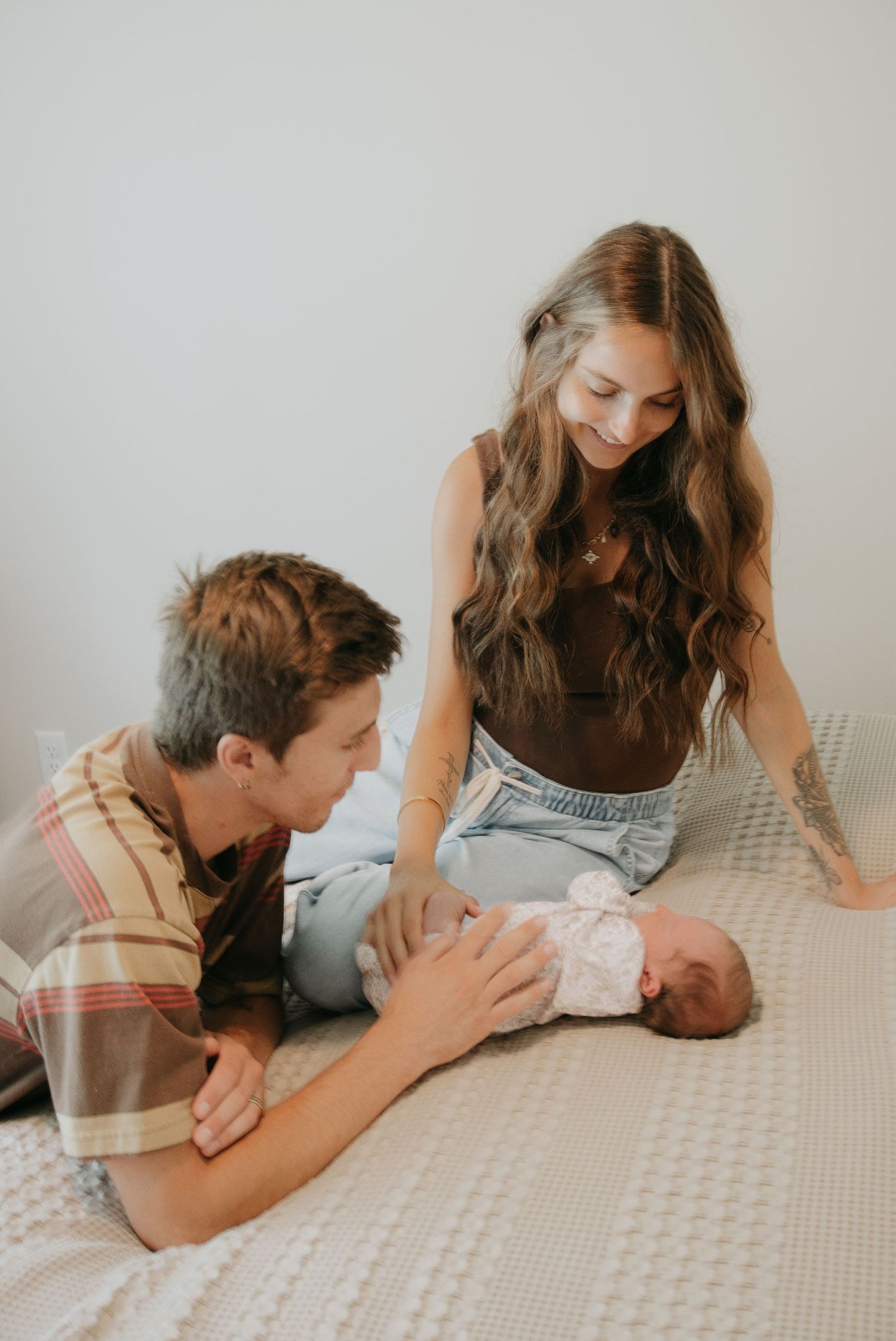 Young couple smiling at their newborn baby while sitting together on a bed with a white knit blanket.
