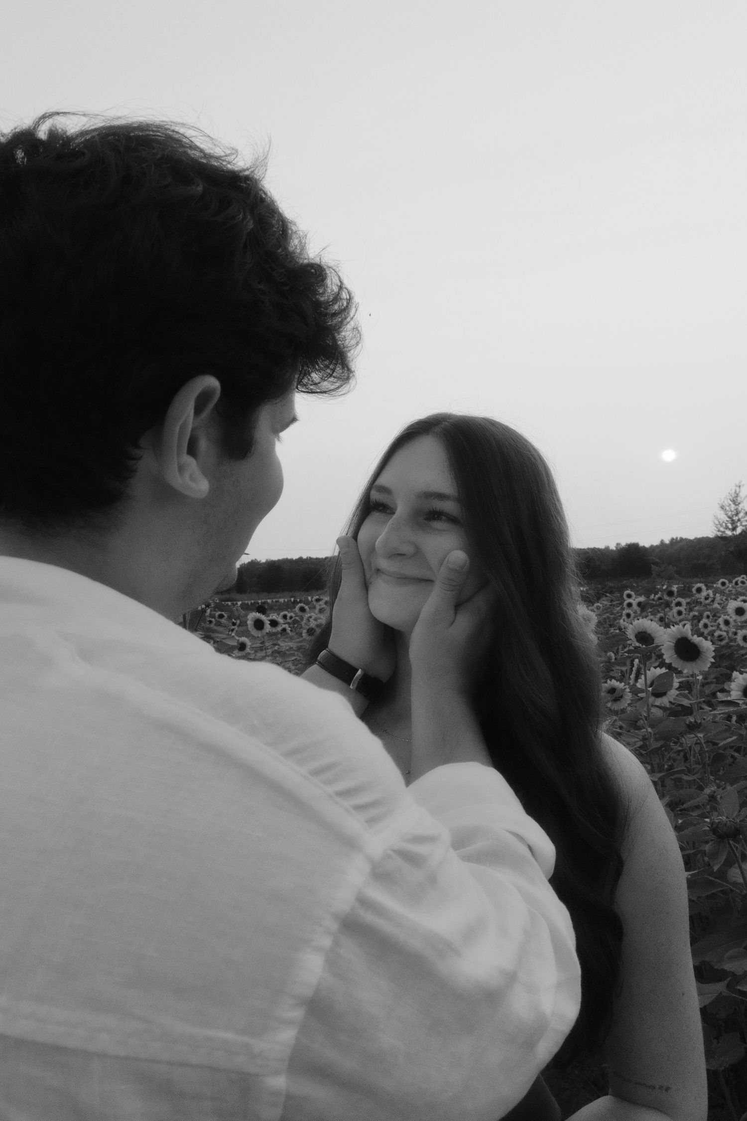 Black and white photo of a man tenderly cupping a smiling woman's face in a sunflower field.