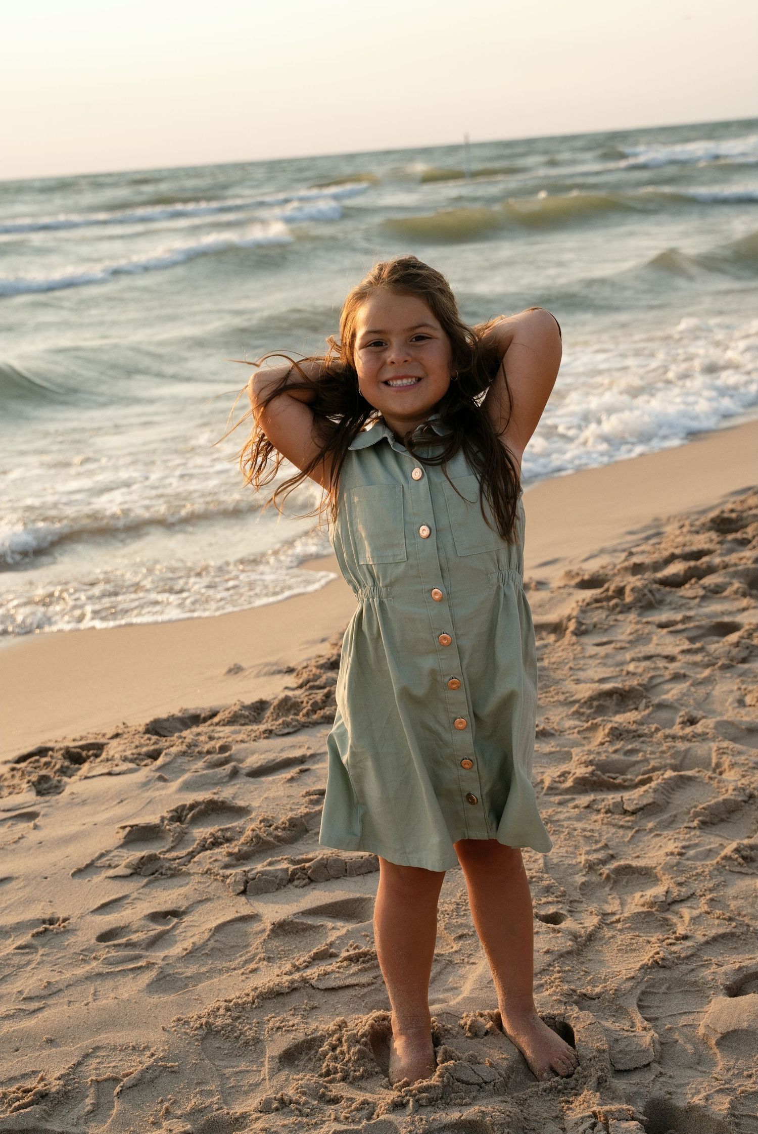 Young girl in a sage green button dress smiling on a sandy beach with ocean waves behind her.