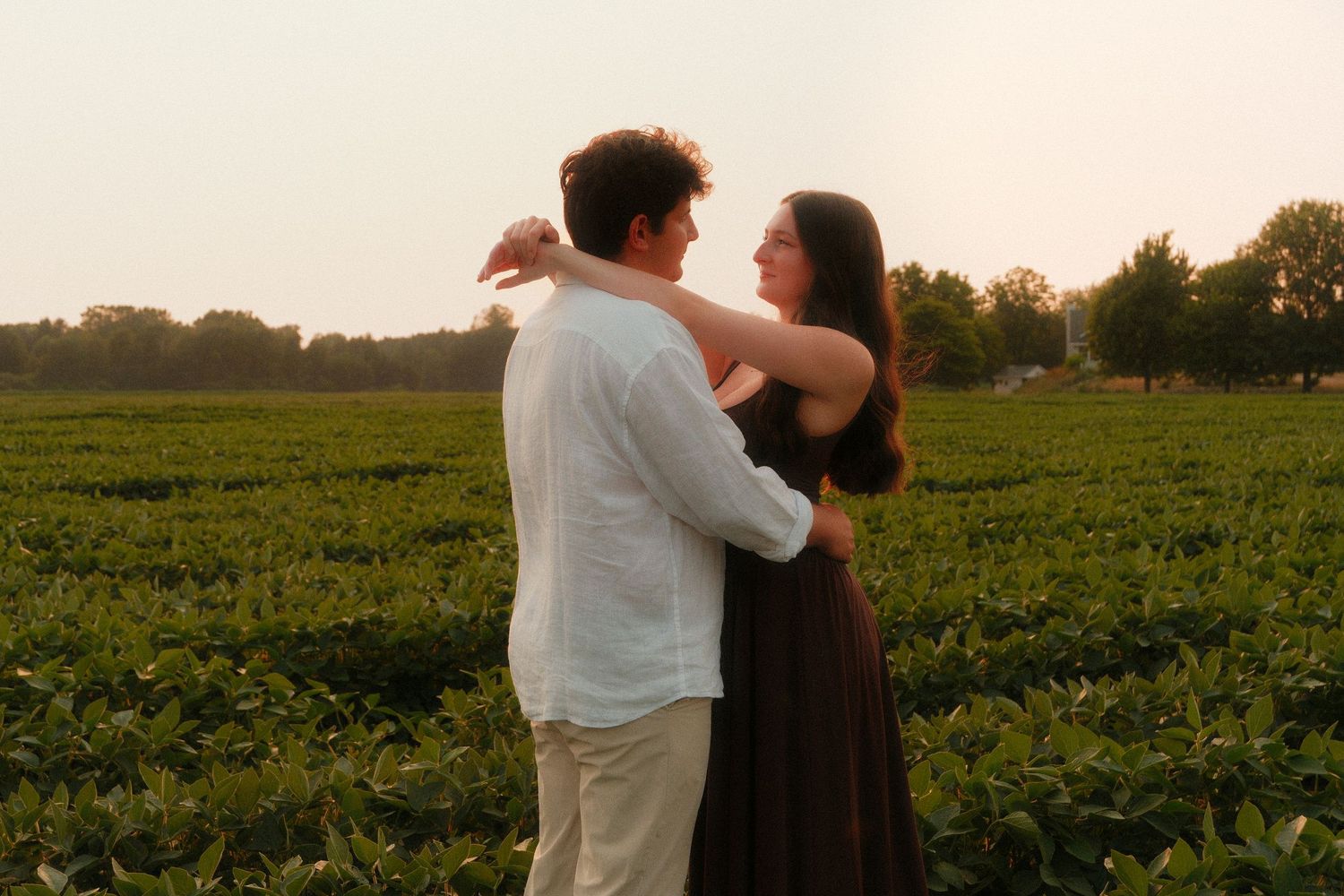 Couple dancing romantically in a lush green farm field at sunset, woman in dark dress, man in white shirt.