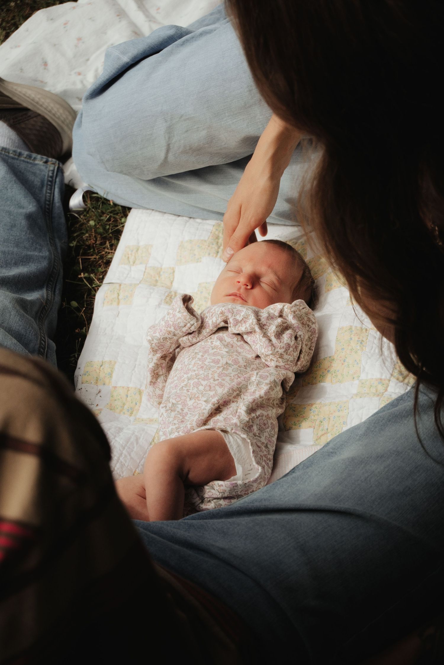 New parents sitting on a vintage quilt outdoors, gently touching their sleeping newborn baby in a floral outfit.