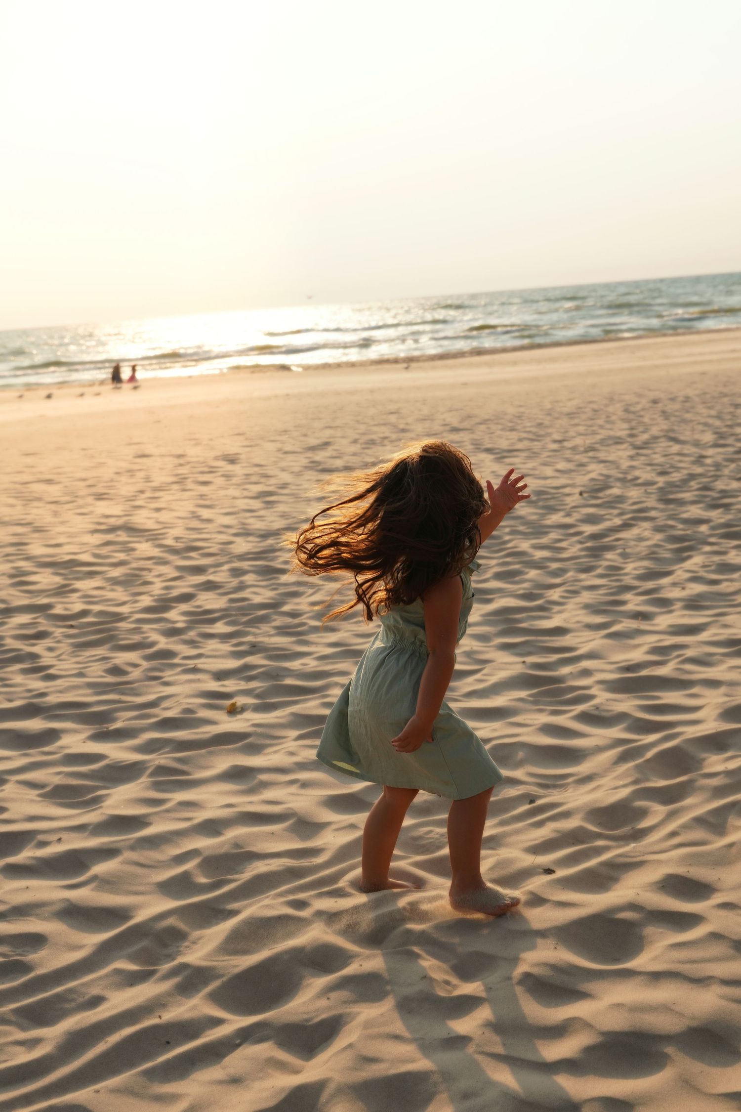 Young girl in green dress dancing barefoot on sandy beach at sunset with ocean waves in background.