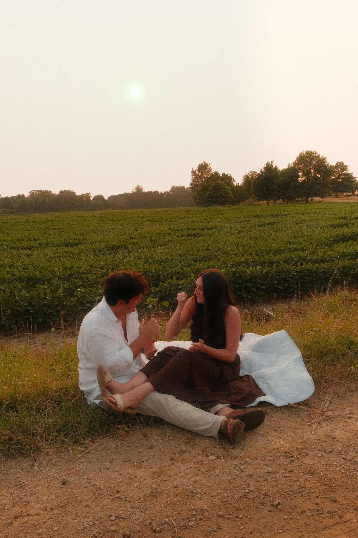 Couple sharing a romantic picnic on a blanket beside lush green fields at golden hour sunset.