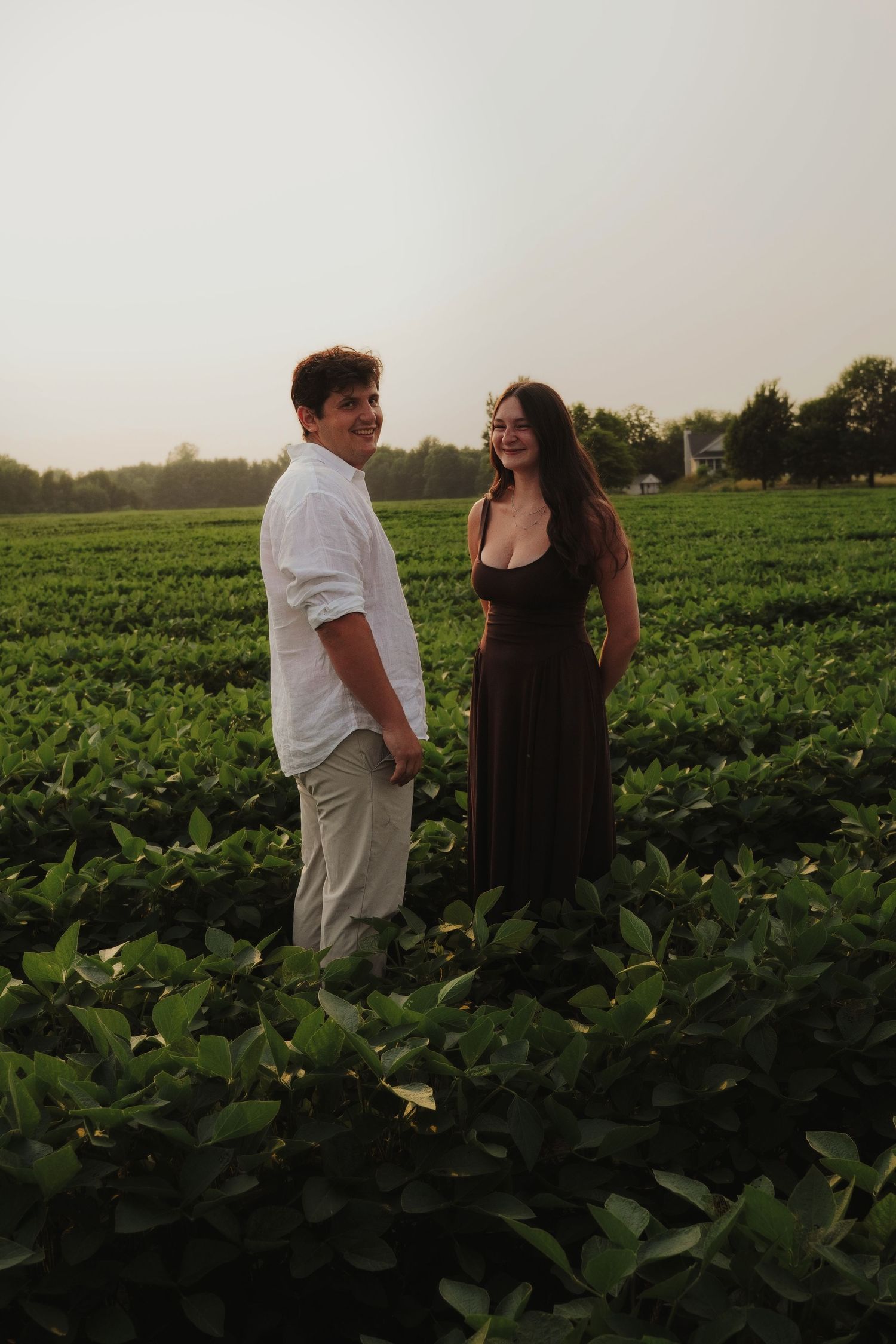 A couple stands smiling in a lush green field at sunset, man in white shirt and woman in dark dress.