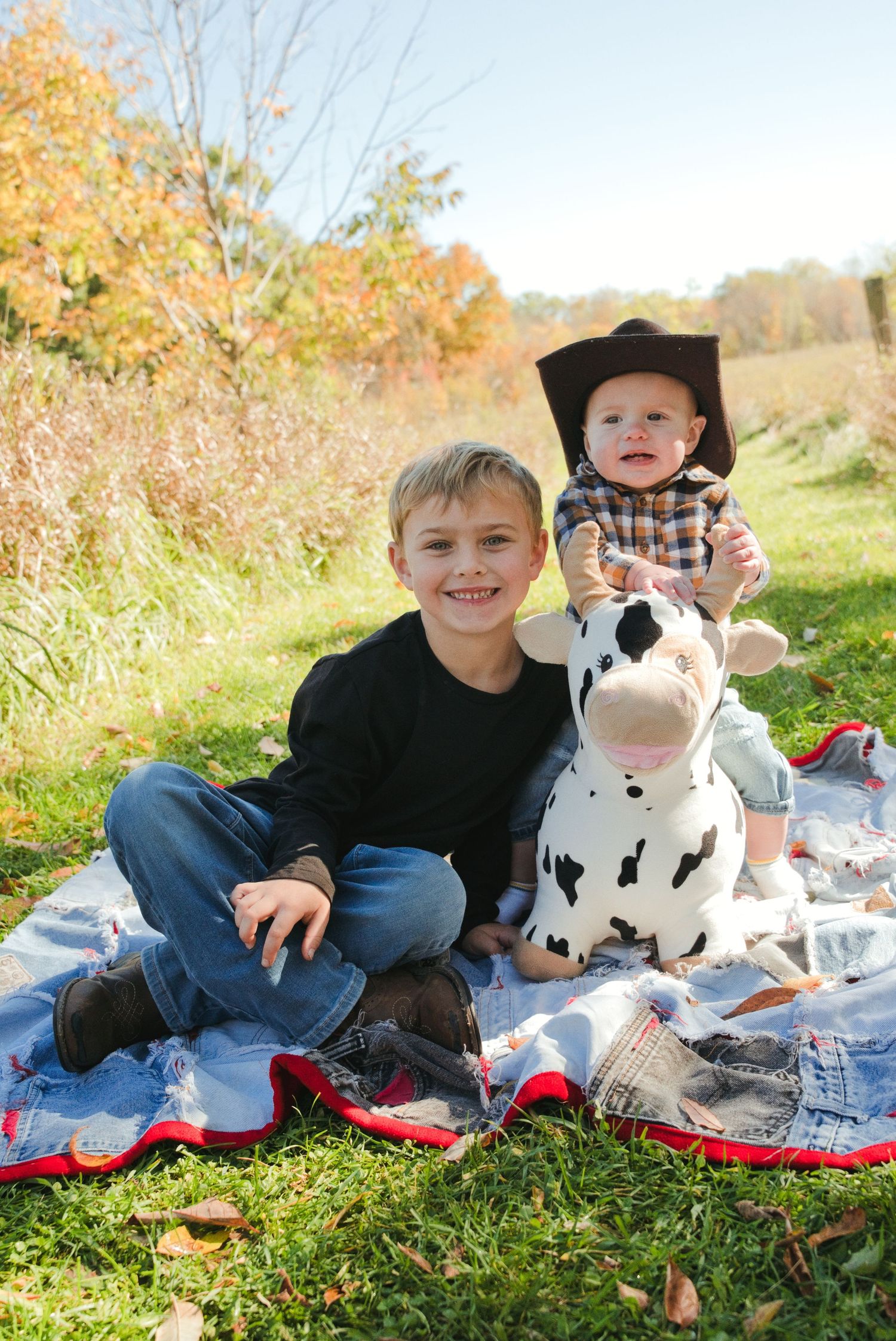 Smiling boy in jeans sits on a quilt beside a baby in a cowboy hat on a toy cow, with autumn trees behind.