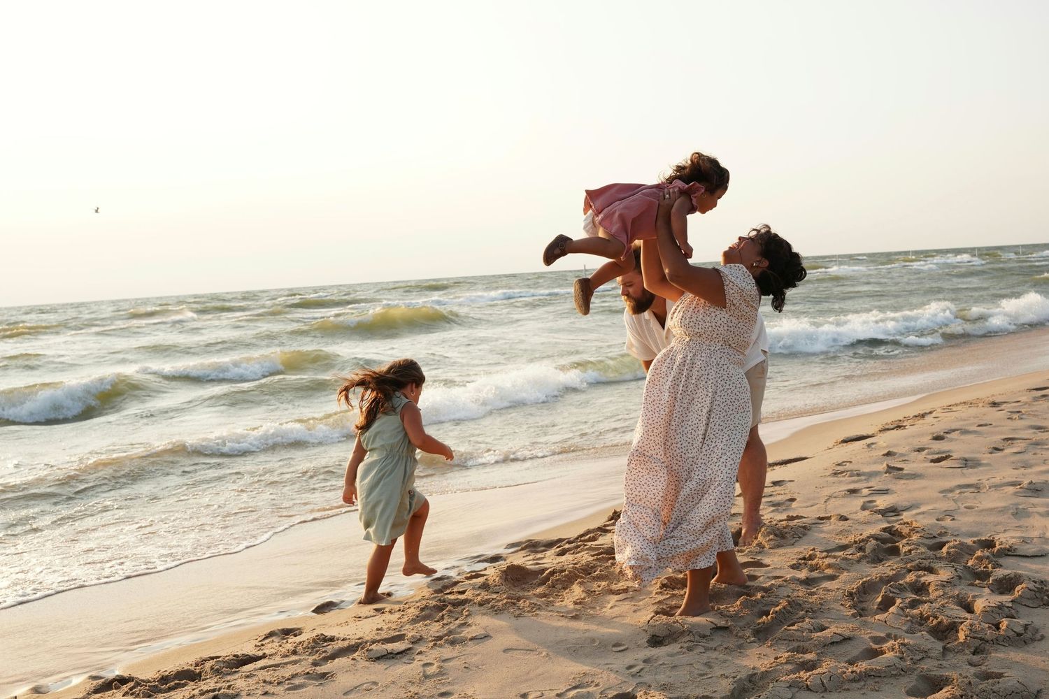 Mother playfully lifting child on sandy beach while another child dances nearby with ocean waves in background.