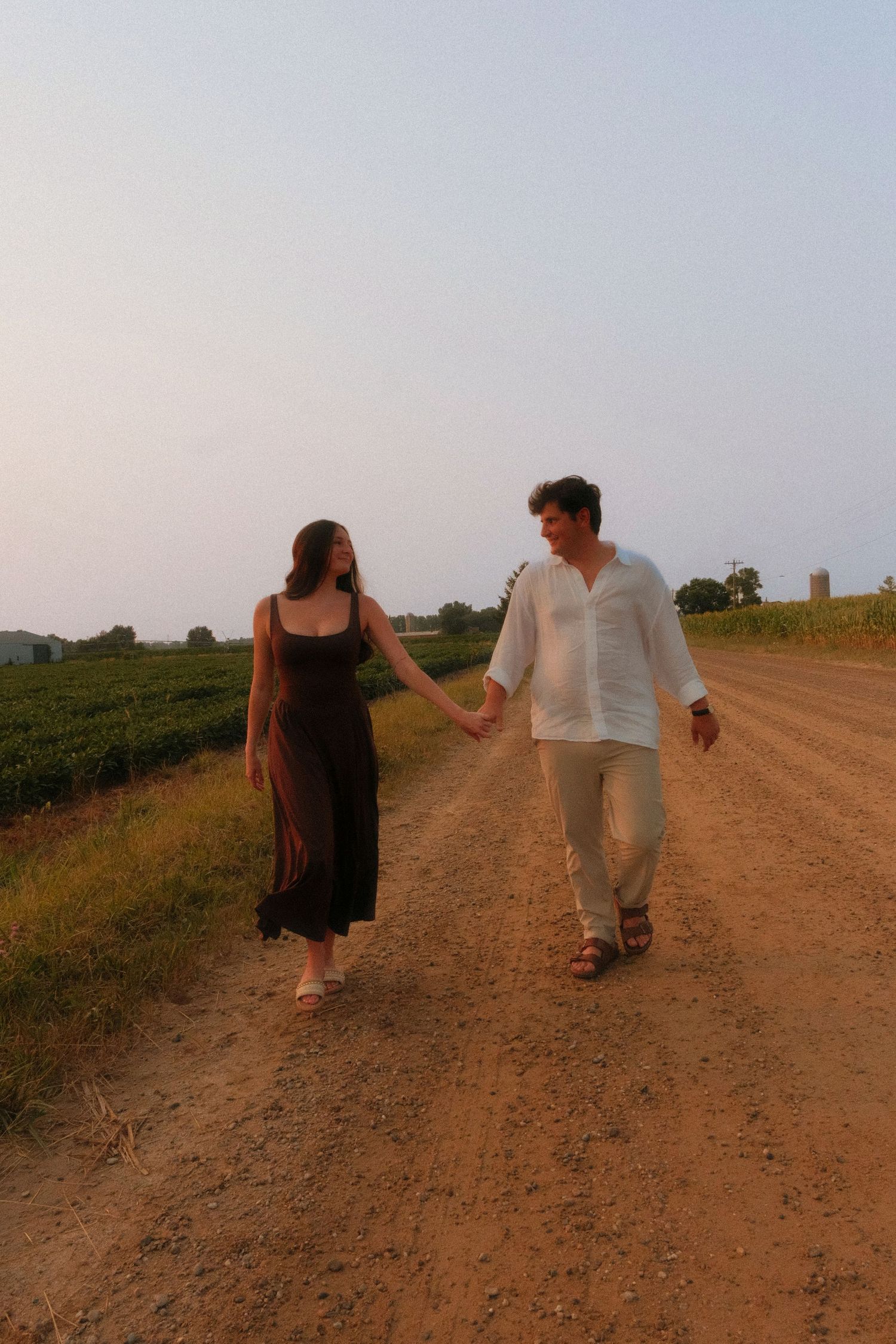 Young couple holding hands strolling down a rustic dirt road surrounded by green fields at sunset.