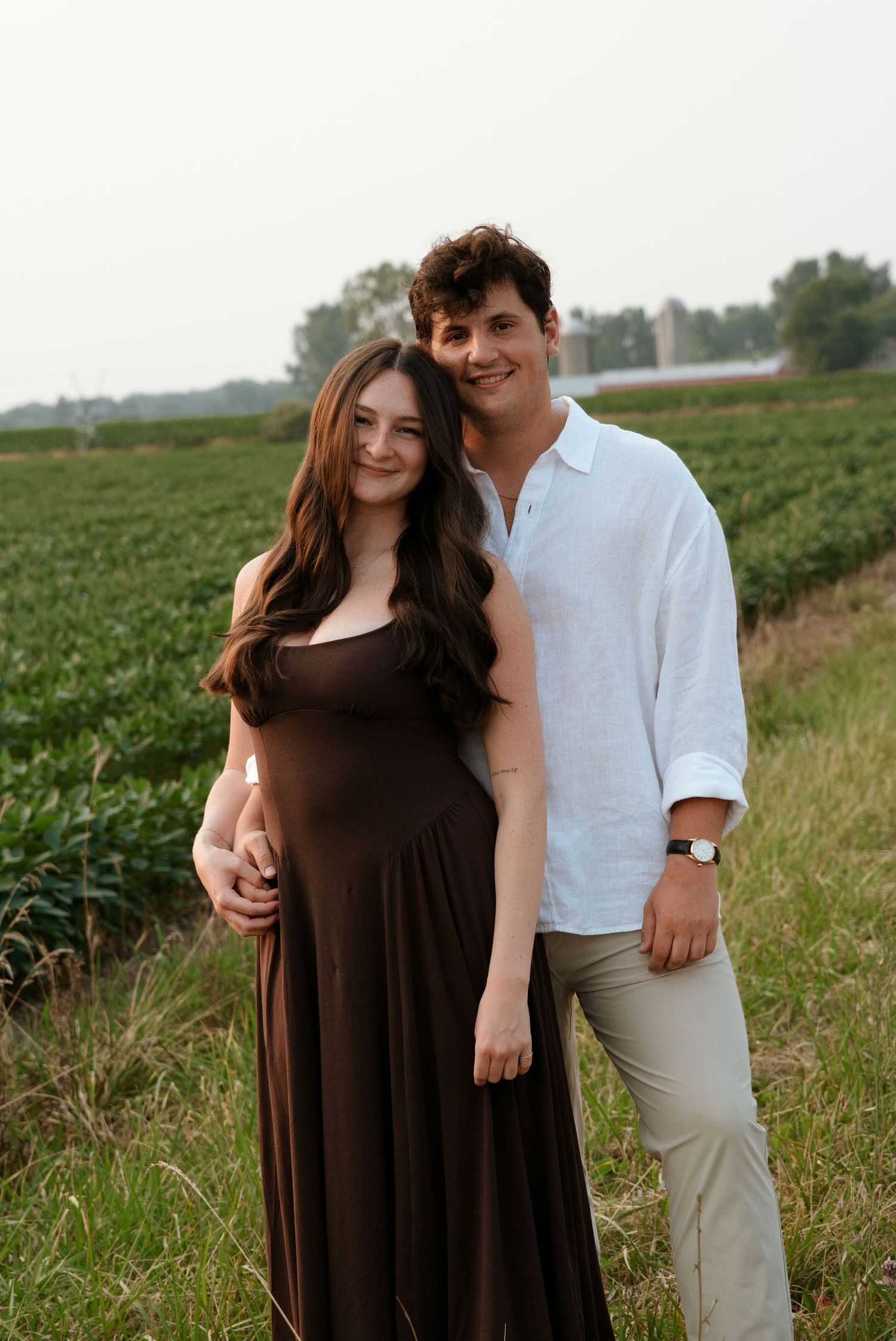 Couple posing in a green field, woman in brown maxi dress with baby bump, man in white linen shirt.