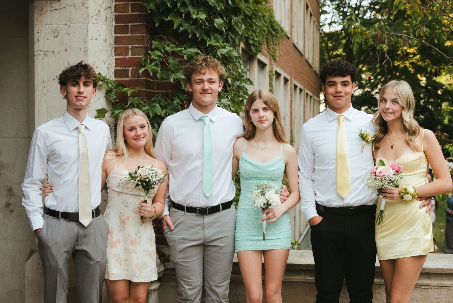 Six teens in semi-formal outfits pose together outdoors beside a brick building, holding small bouquets, with boys in ties and girls in past