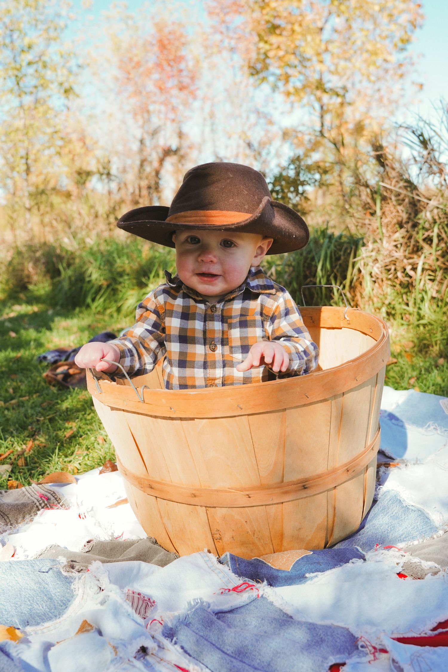 Smiling baby in a brown cowboy hat and plaid shirt sits inside a wooden apple basket on a quilt with autumn trees behind.