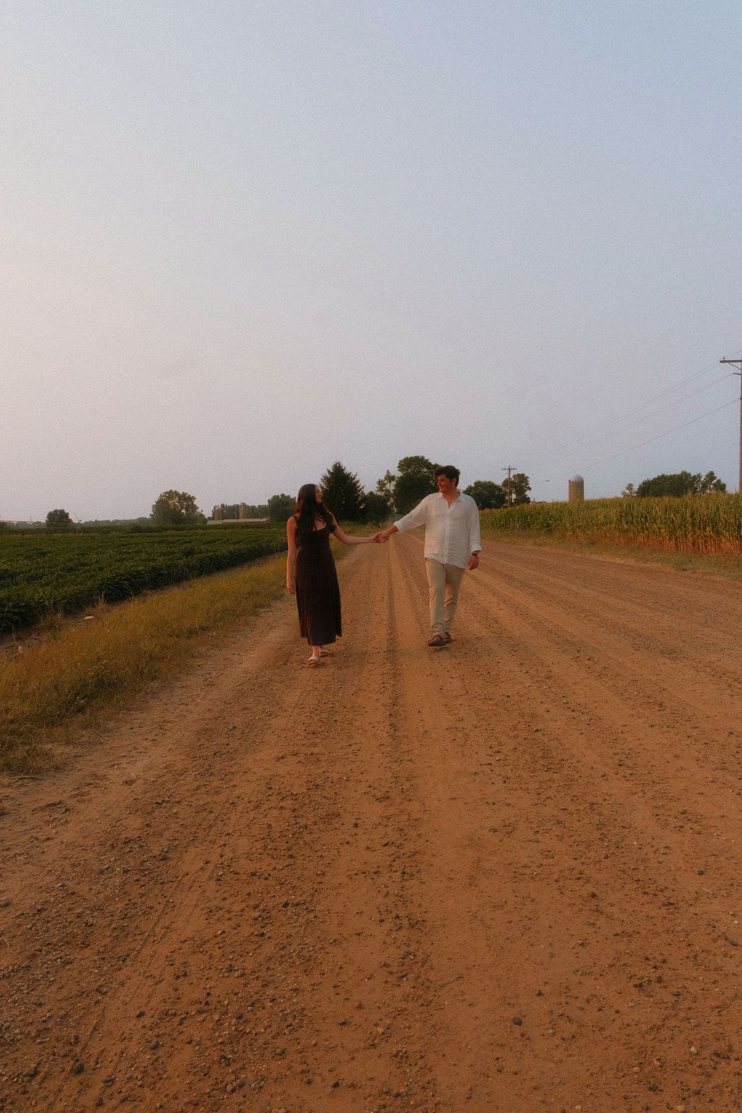 Couple holding hands while walking down a rural dirt road surrounded by green farmland at dusk.
