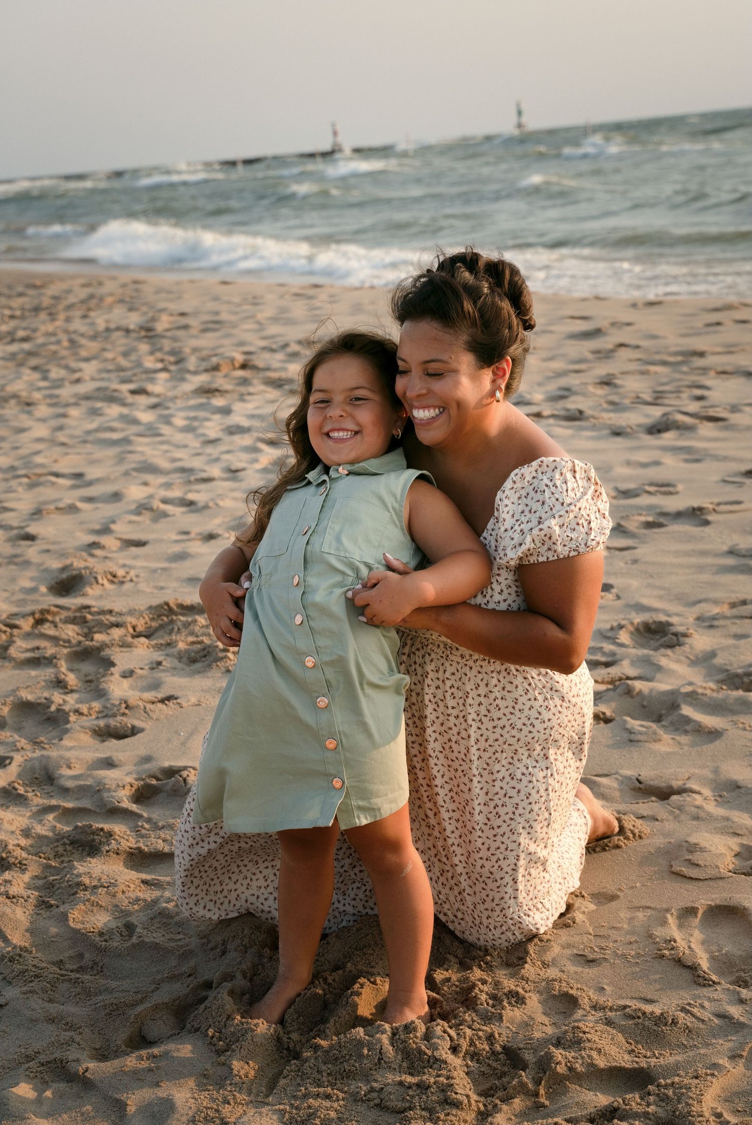 A woman kneeling on sandy beach hugging a smiling young girl in a green dress, with ocean waves in the background.