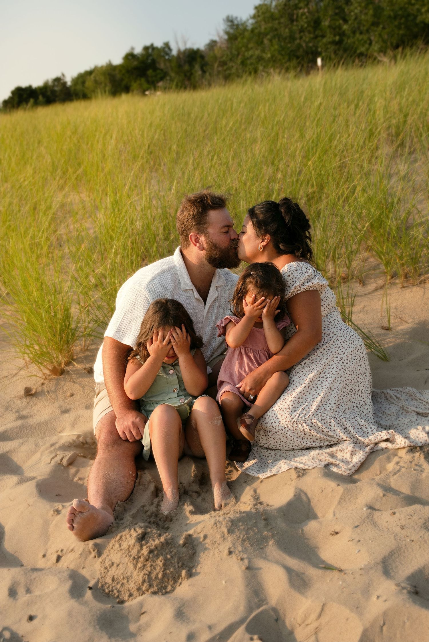 A family sits on sandy beach dunes, parents kissing while two young children sit between them, grass in background.