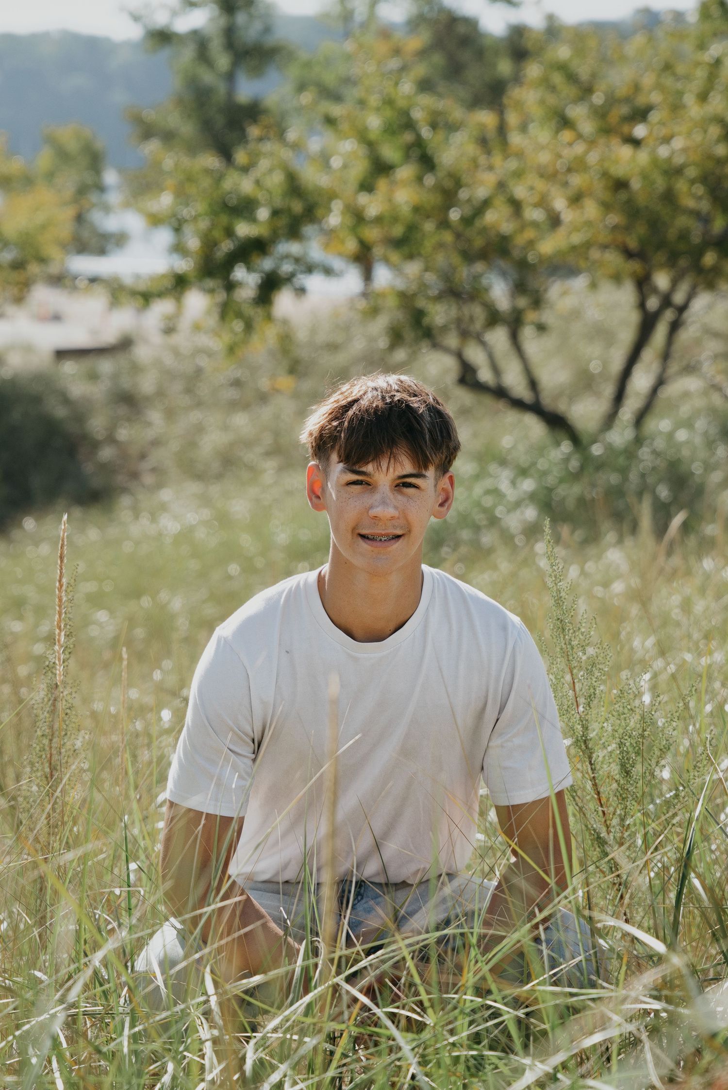 A smiling teenage boy with braces sits in a lush green meadow wearing a white t-shirt and denim shorts on a sunny day.