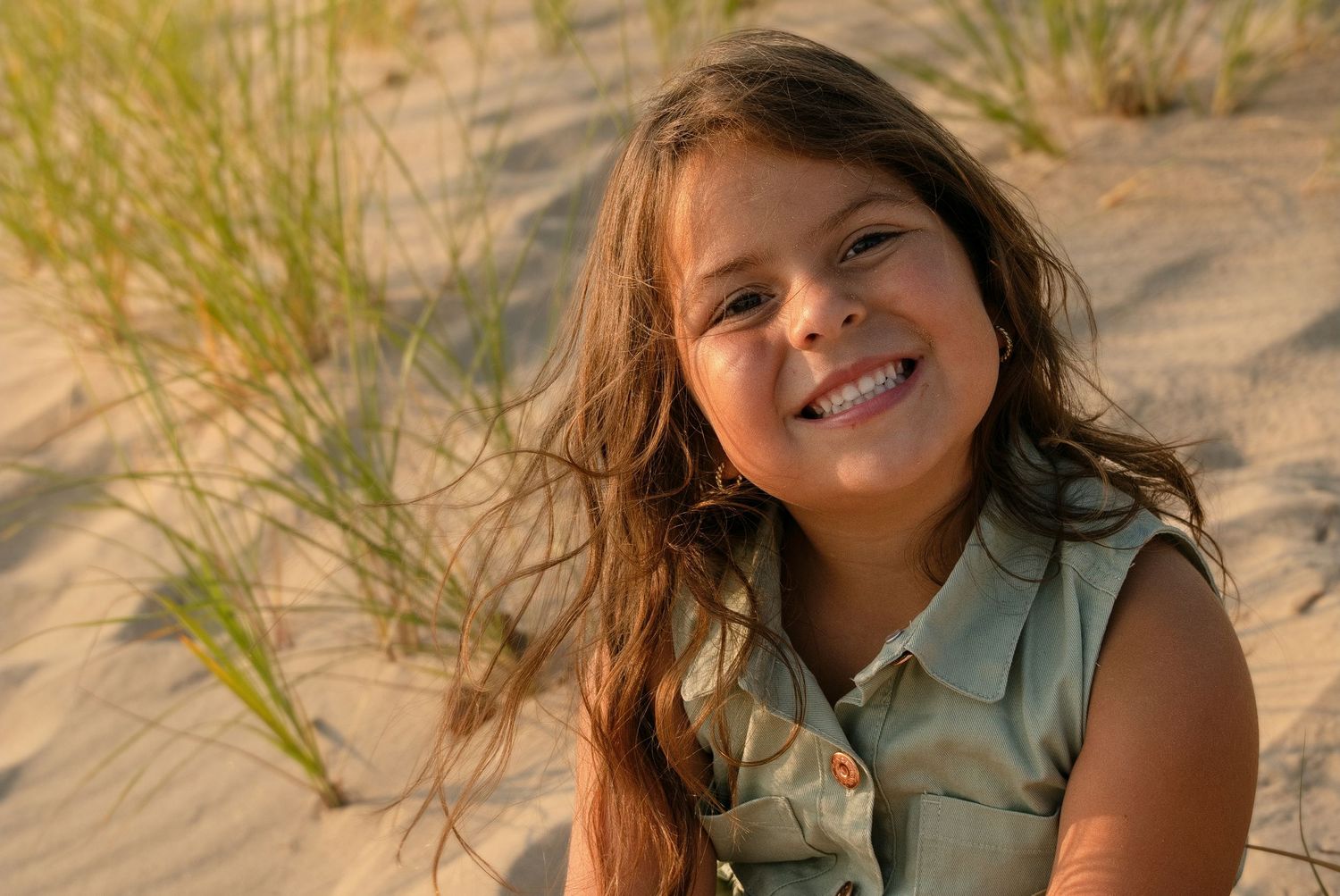 A smiling young girl with long brown hair sits on sandy beach dunes with green grass in the background.