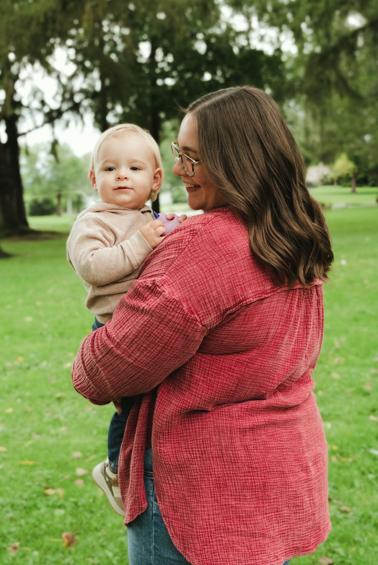 Smiling woman in glasses and a red linen shirt holds a baby in a beige sweater while standing on a green lawn in a leafy park.