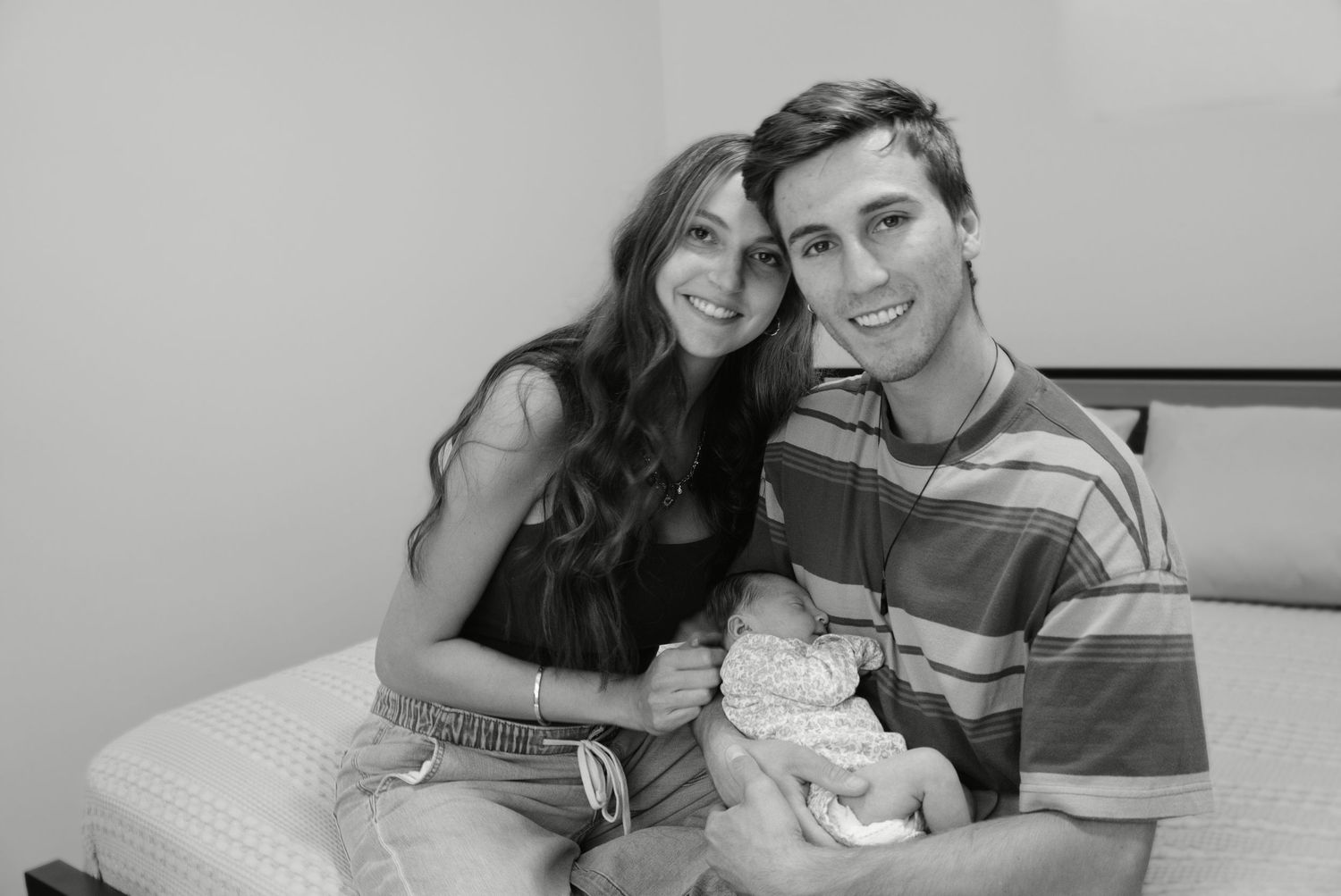 Young couple smiling on bed holding newborn baby in black and white photo.
