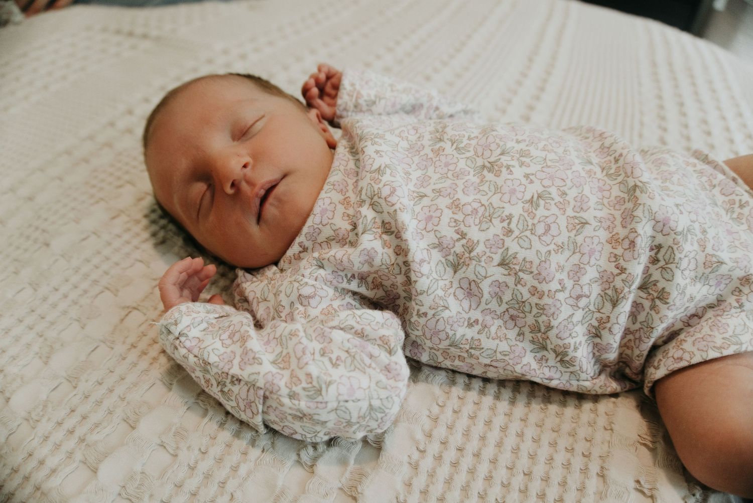 Peaceful newborn baby sleeping on a white textured blanket wearing a soft floral onesie.