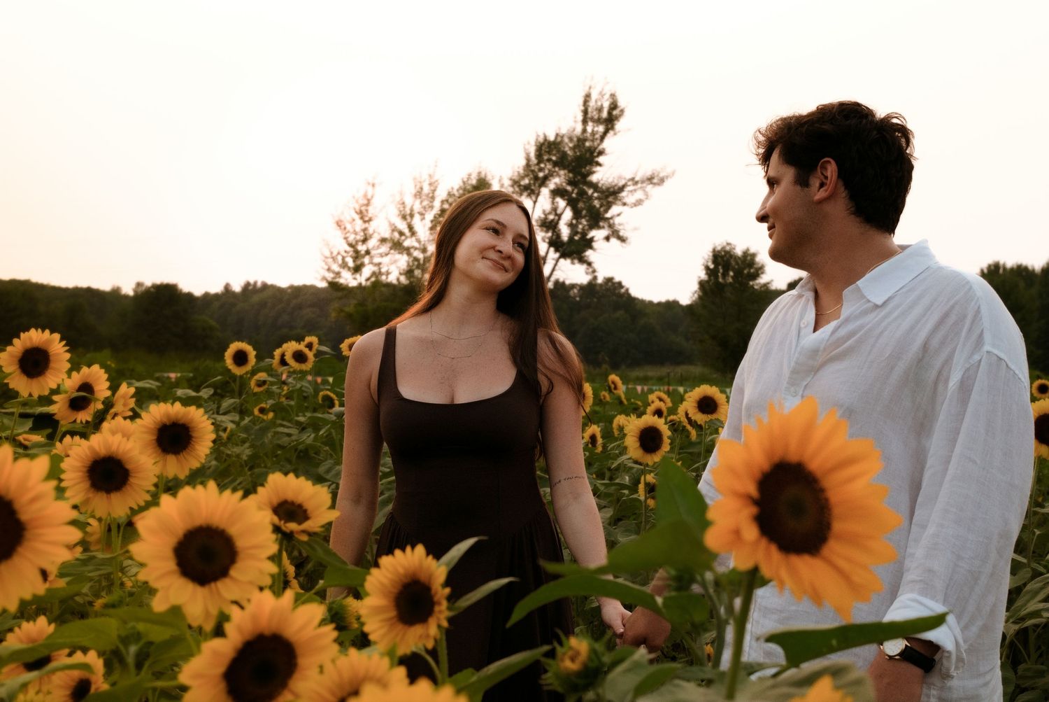 Couple smiling at each other in a vibrant sunflower field during sunset, woman in black dress, man in white shirt.