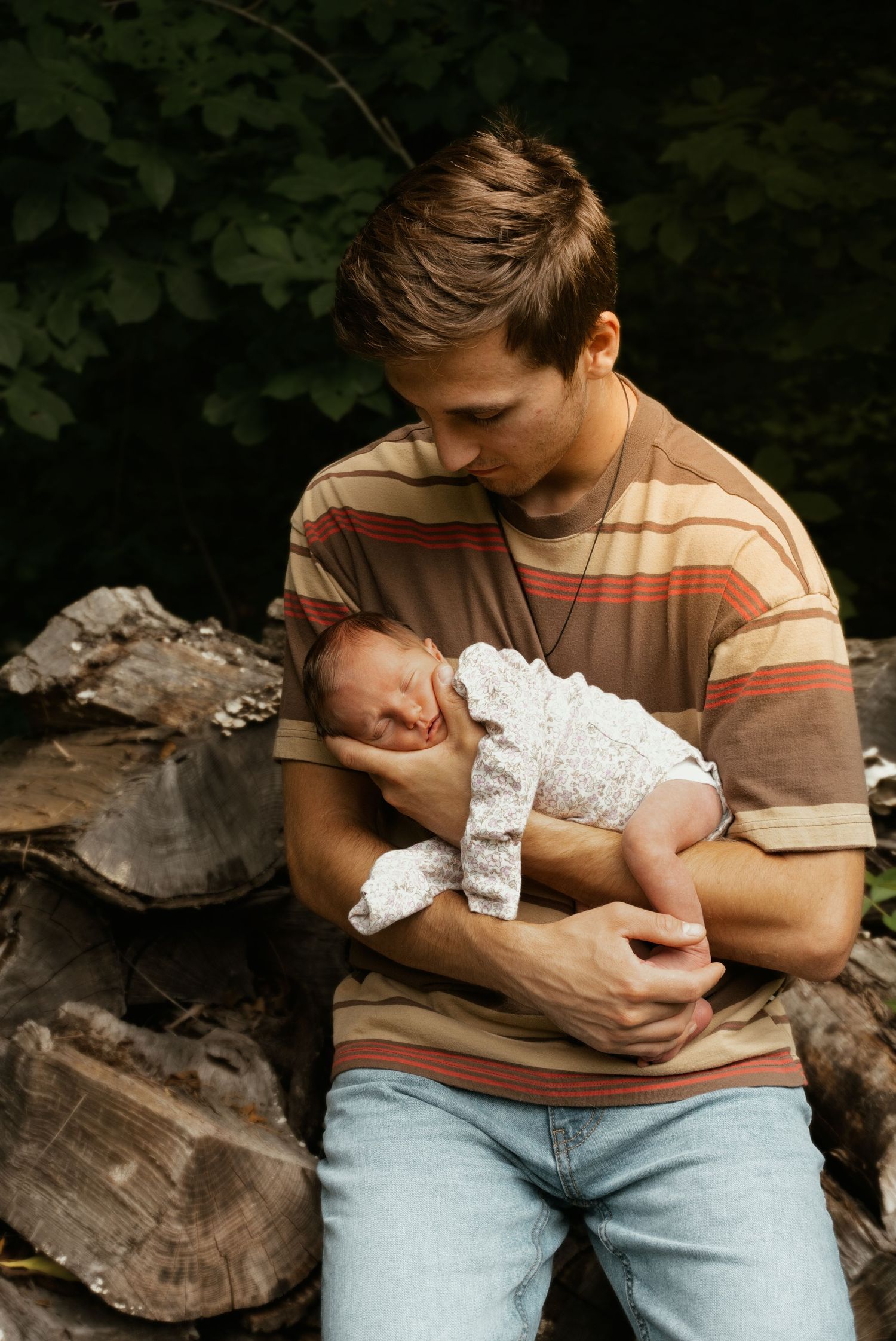 Young father in striped shirt tenderly cradling sleeping newborn baby outdoors near stacked logs.