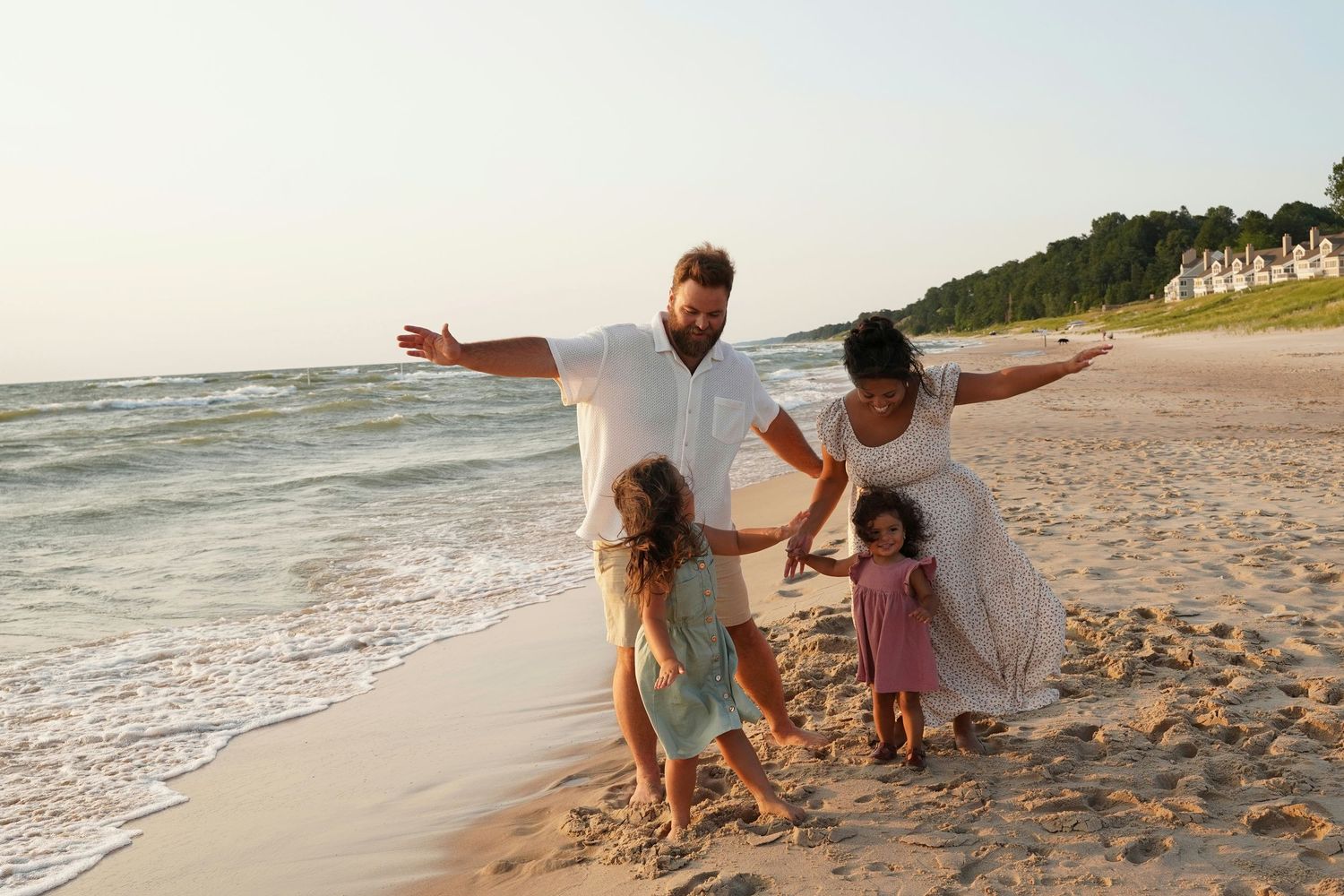 Family of four joyfully playing on a sandy beach at sunset, with parents swinging two young daughters near the ocean waves.